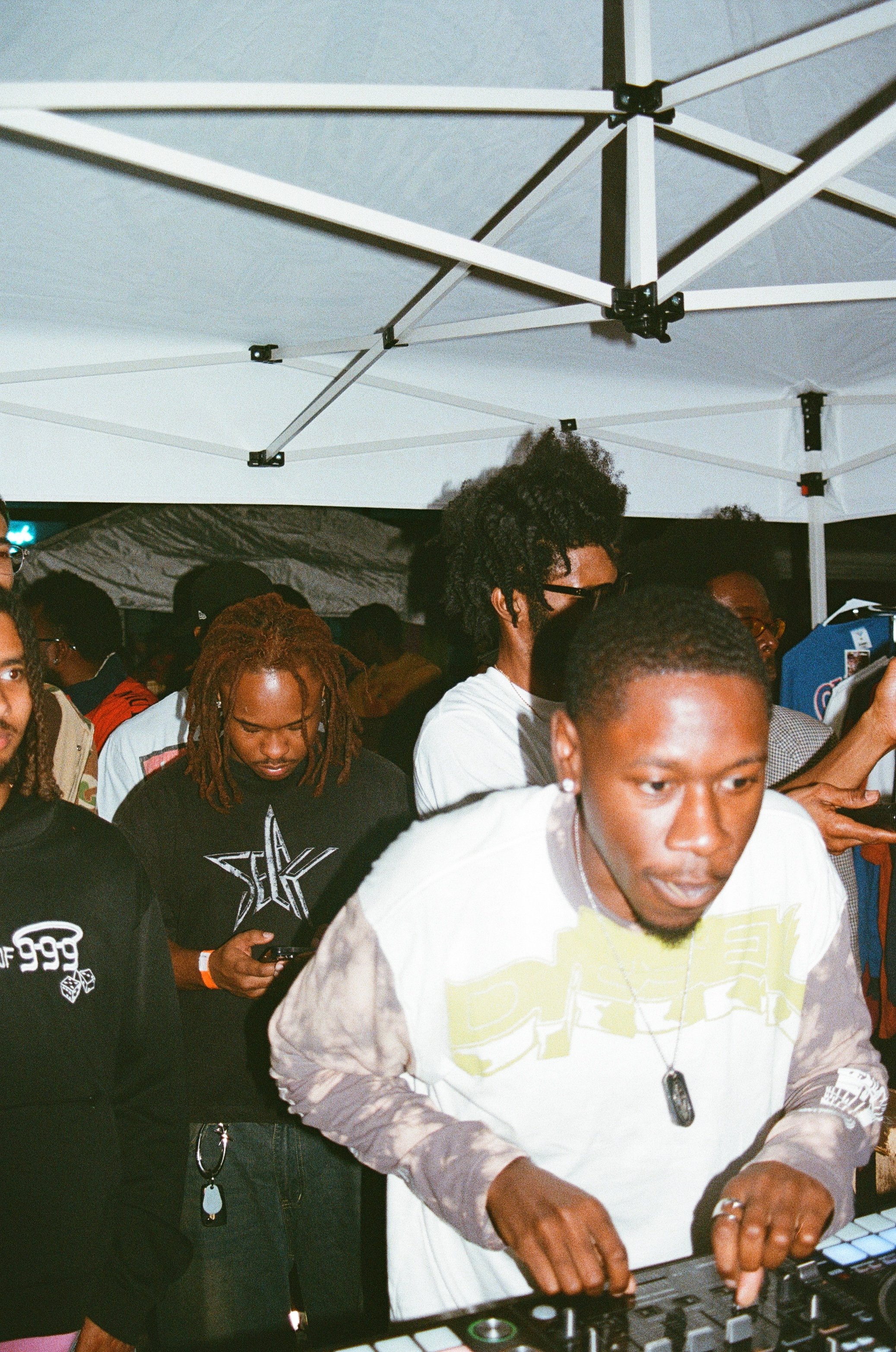 A group of people at an outdoor event, with a DJ using a mixing console under a white canopy tent at night.