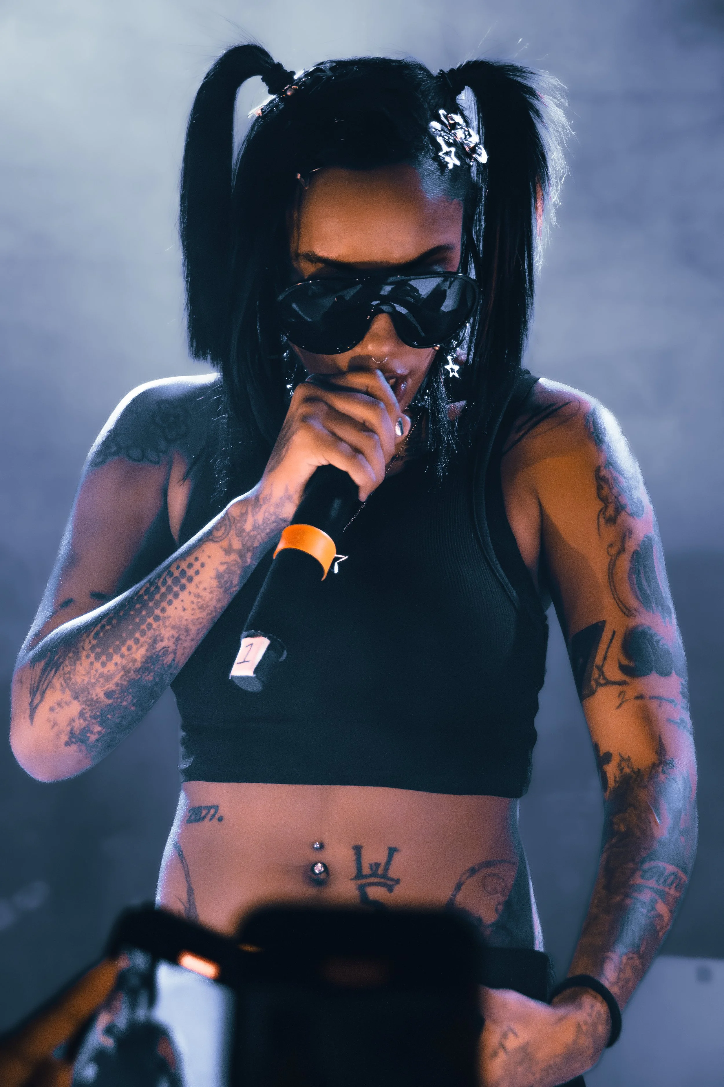 Tattoed female performer with black hair in pigtails, wearing sunglasses and a black crop top, holding a microphone during a performance.