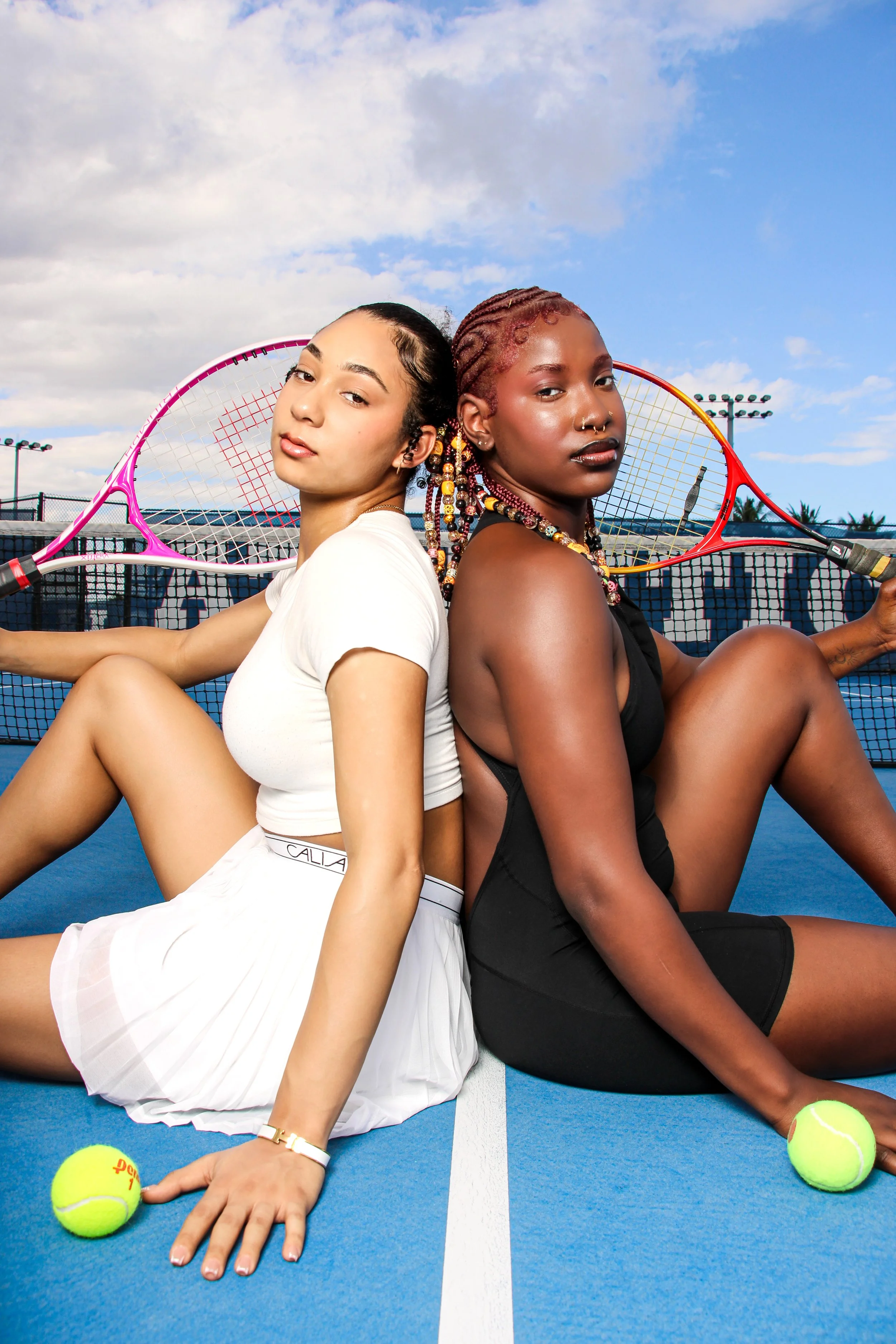 Two young women sitting back-to-back on a tennis court with tennis rackets and tennis balls, under a partly cloudy sky.