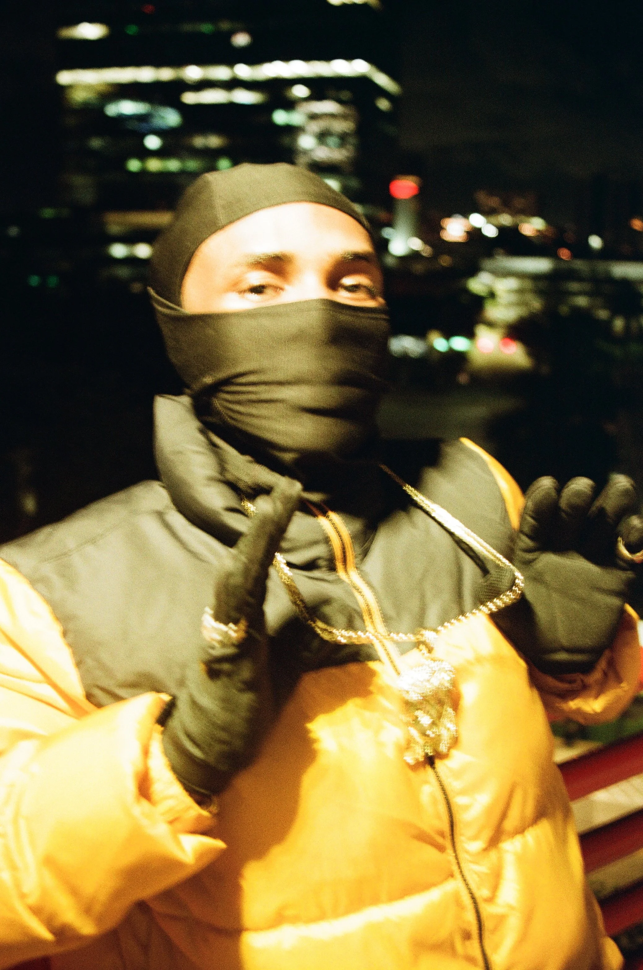 A person dressed in a black face mask, black hat, orange and black jacket, and leather gloves, standing outdoors at night with city lights and buildings in the background.