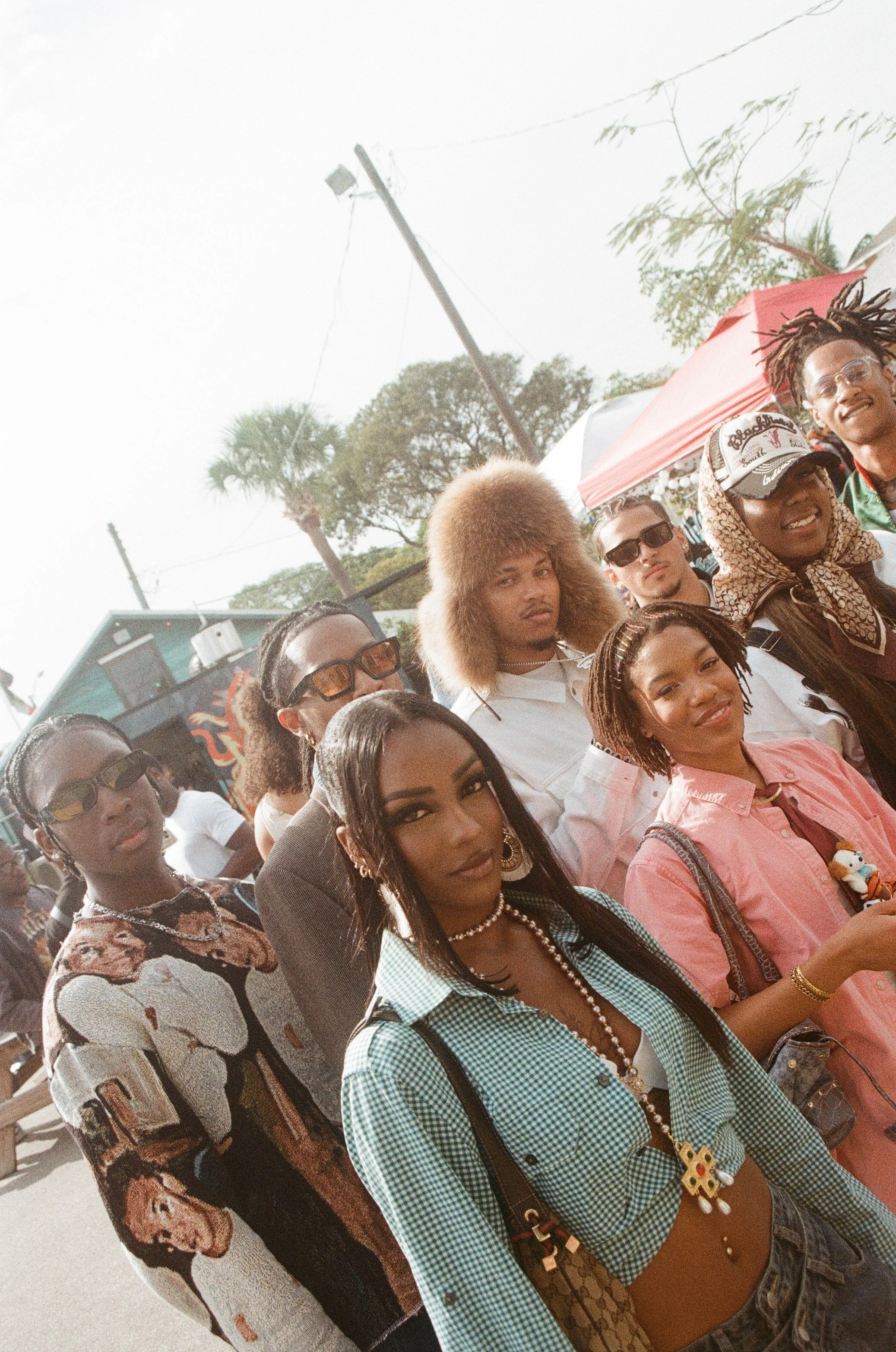Group of diverse people gathered outdoors, some wearing sunglasses, a woman in a patterned jacket, a woman in a pink shirt holding a toy, a man with a fluffy hairpiece, and another person with dreadlocks, against tents and trees.