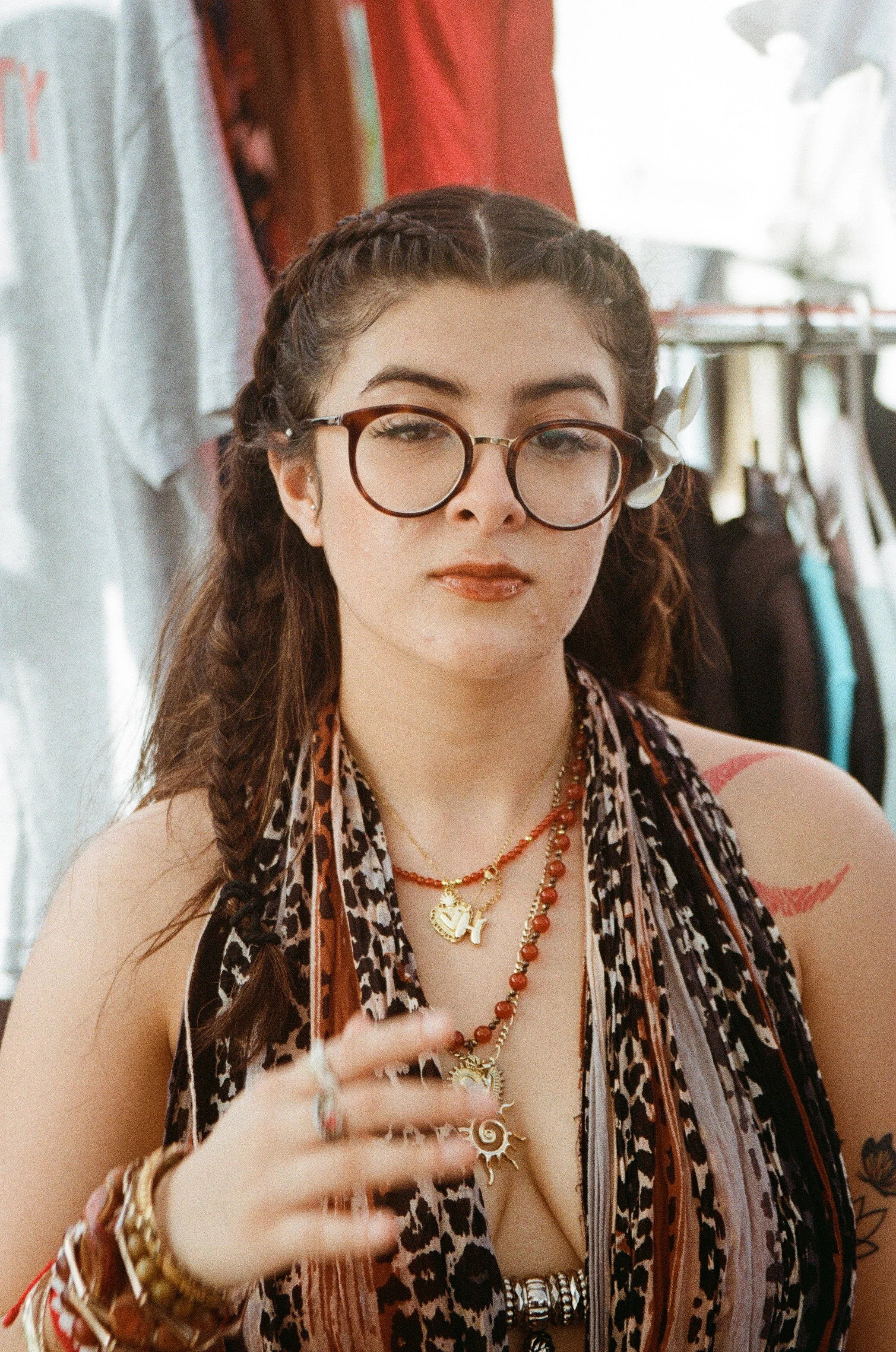 A young woman with glasses, braided hair, and a scarf, wearing multiple necklaces and bracelets, in a clothing or accessories store.