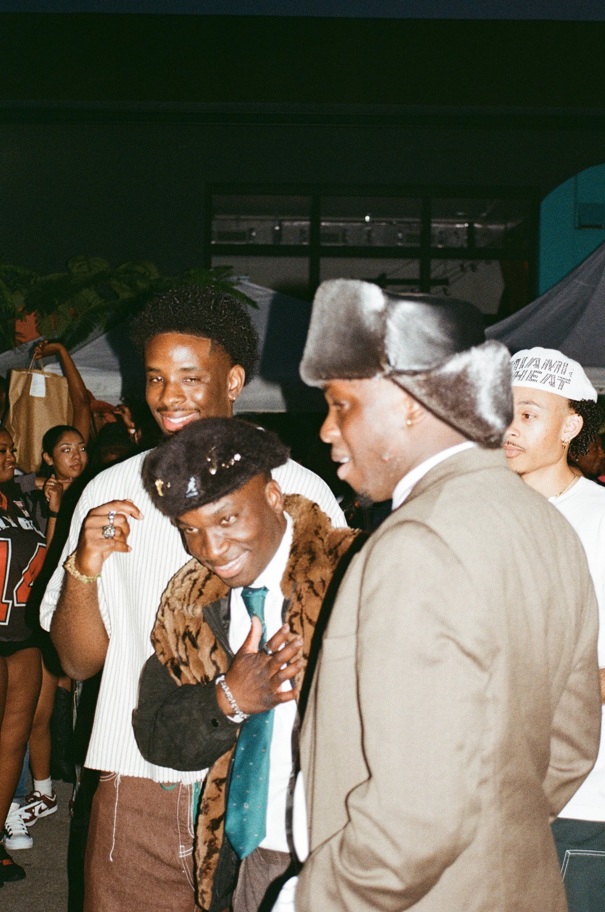 A group of young men at an indoor event, dressed in trendy outfits, engaging in conversation and smiling, with a crowd in the background.