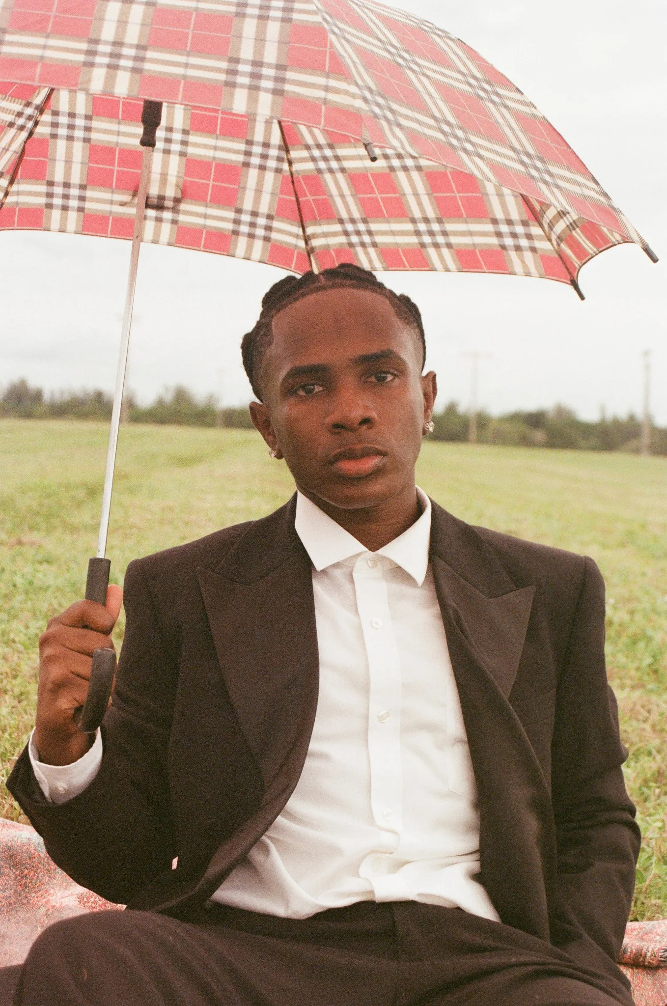 A young man in a tuxedo, holding a patterned umbrella, sitting outdoors on a cloudy day.
