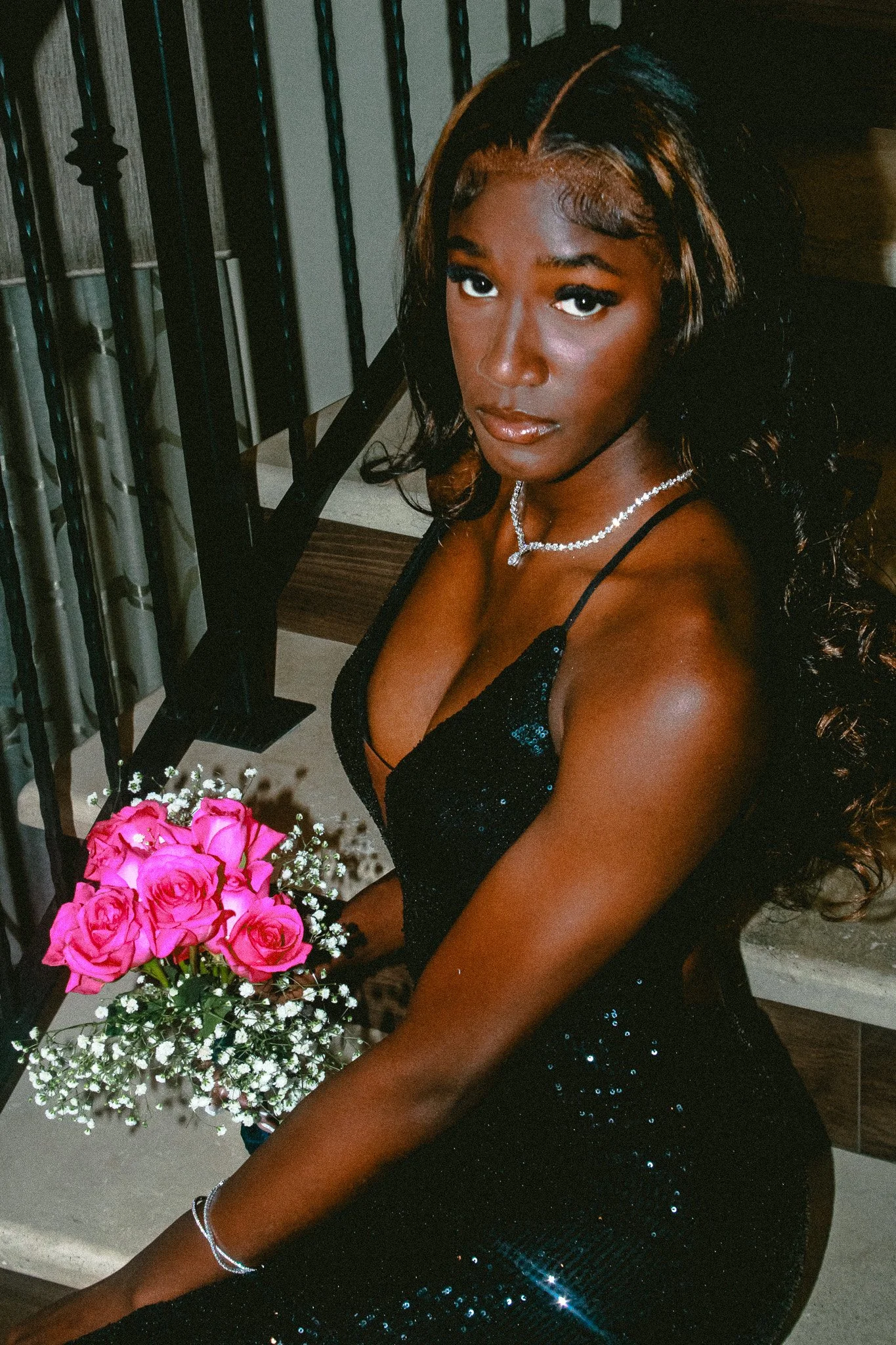 A woman in a black sequined dress holding a bouquet of pink roses and white baby's breath, sitting on stairs, wearing a pearl necklace and bracelet, with long wavy hair.