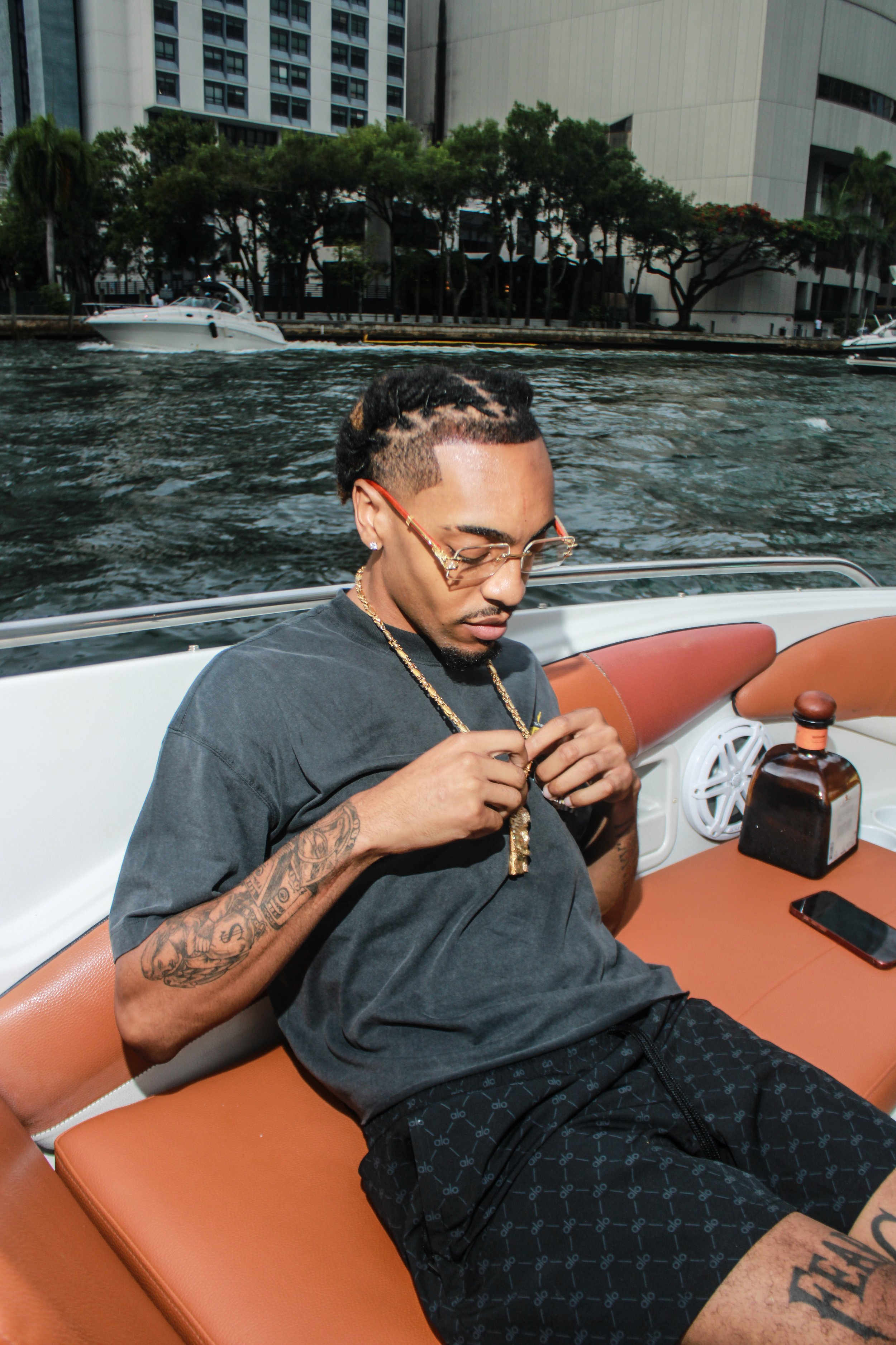 Man with tattoos and glasses sitting on a boat, holding a gold chain, with water and city buildings in the background.