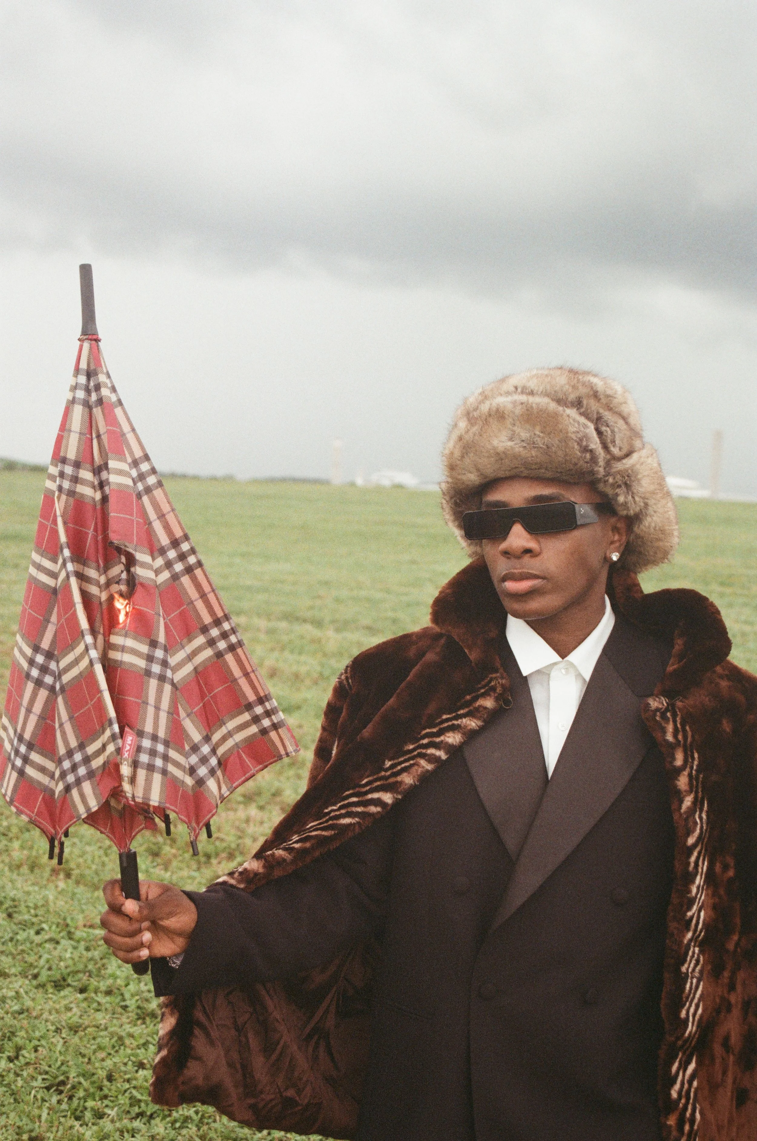 Person wearing a fur hat, sunglasses, and a fur coat, holding a plaid umbrella outdoors on a cloudy day.
