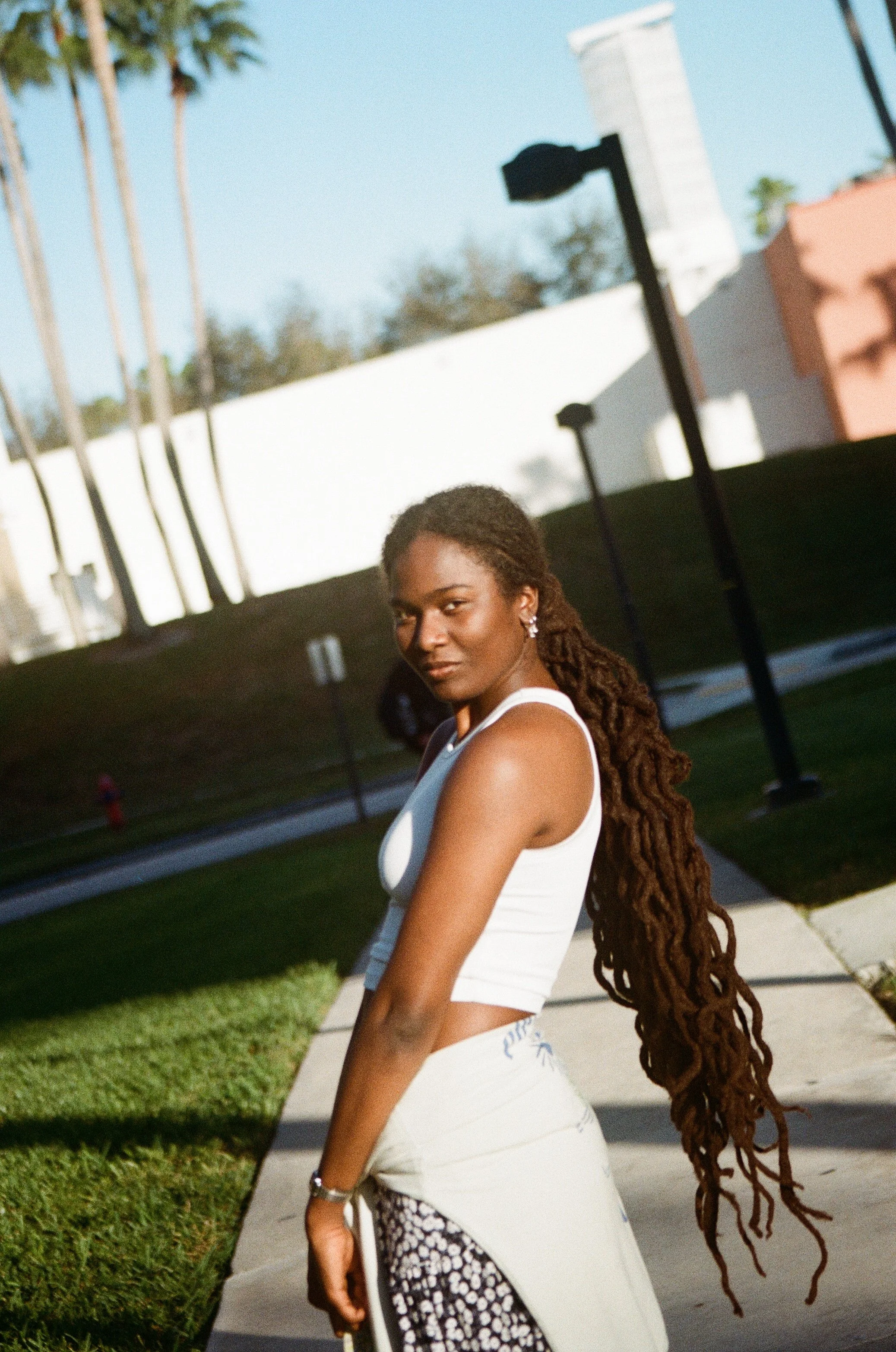 A young woman with long dreadlocks wearing a white tank top and patterned pants standing on a sidewalk in a sunny outdoor setting.