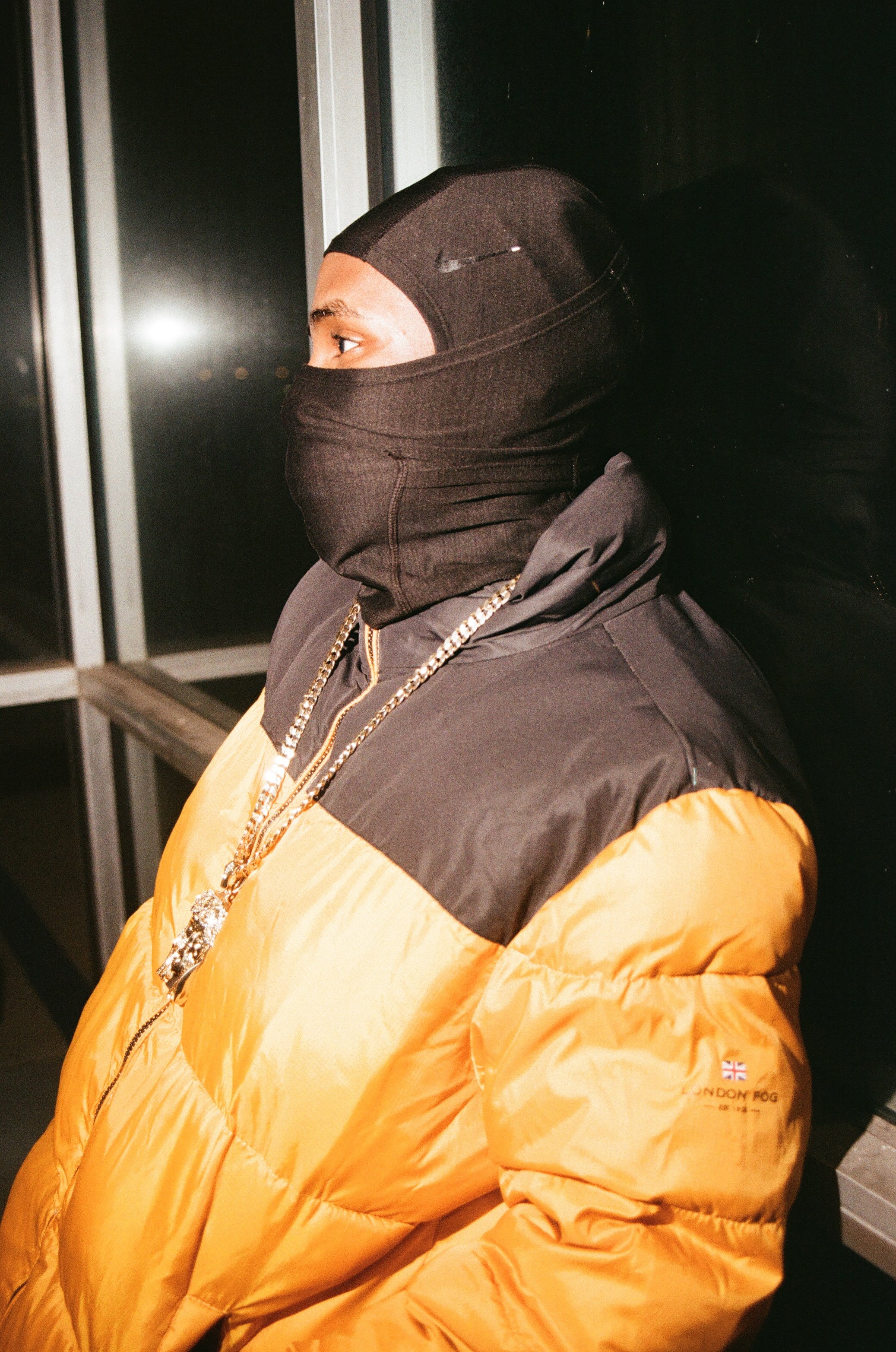 A person wearing a black ski mask and a yellow and black puffer jacket, with gold chains around their neck, standing in front of a dark background.