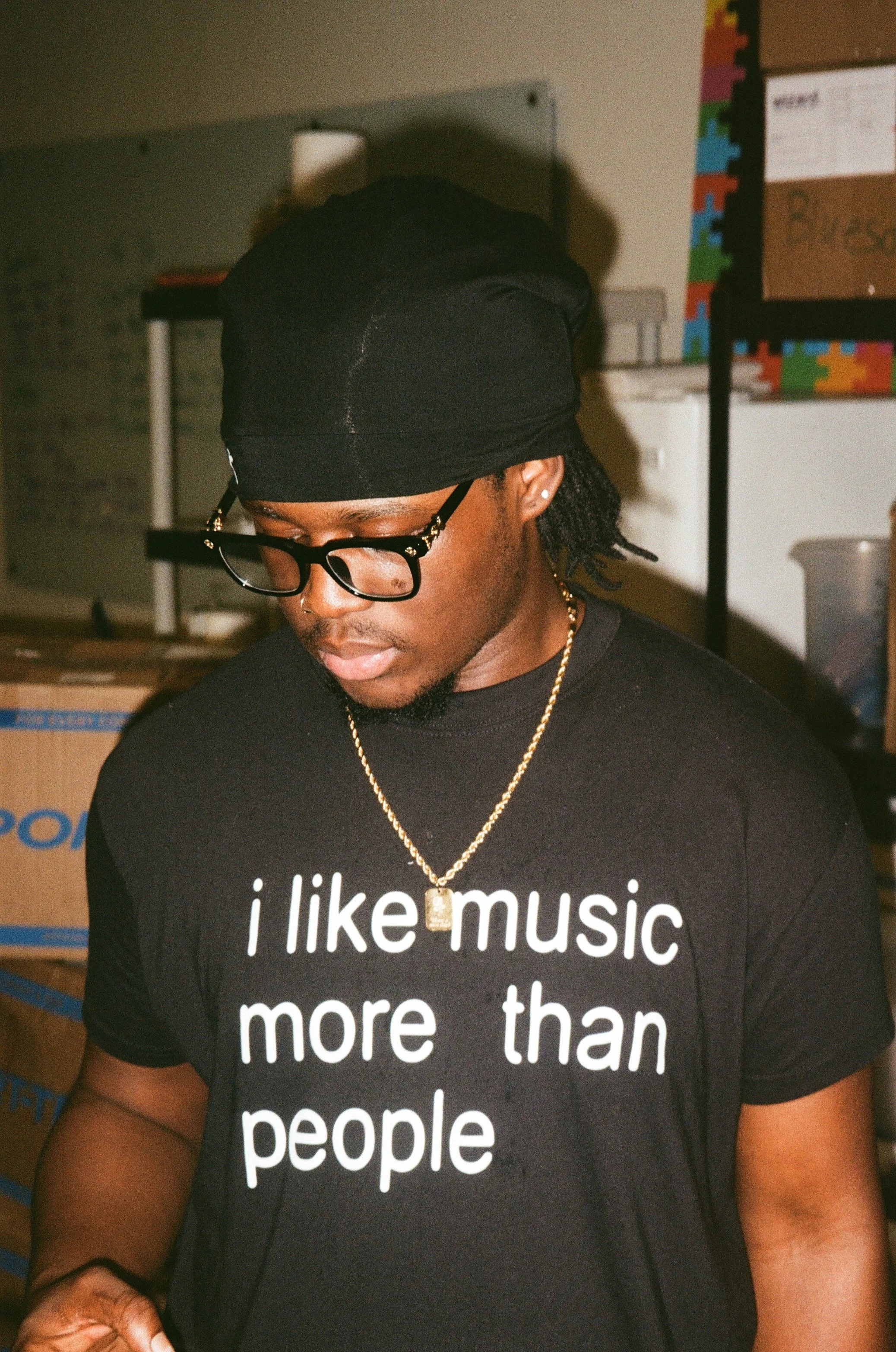 A young man wearing a black durag, glasses, and a black t-shirt with white text reading 'I like music more than people'; he is also wearing a gold necklace with a pendant. He is looking down, with a blurred background that includes cardboard boxes an