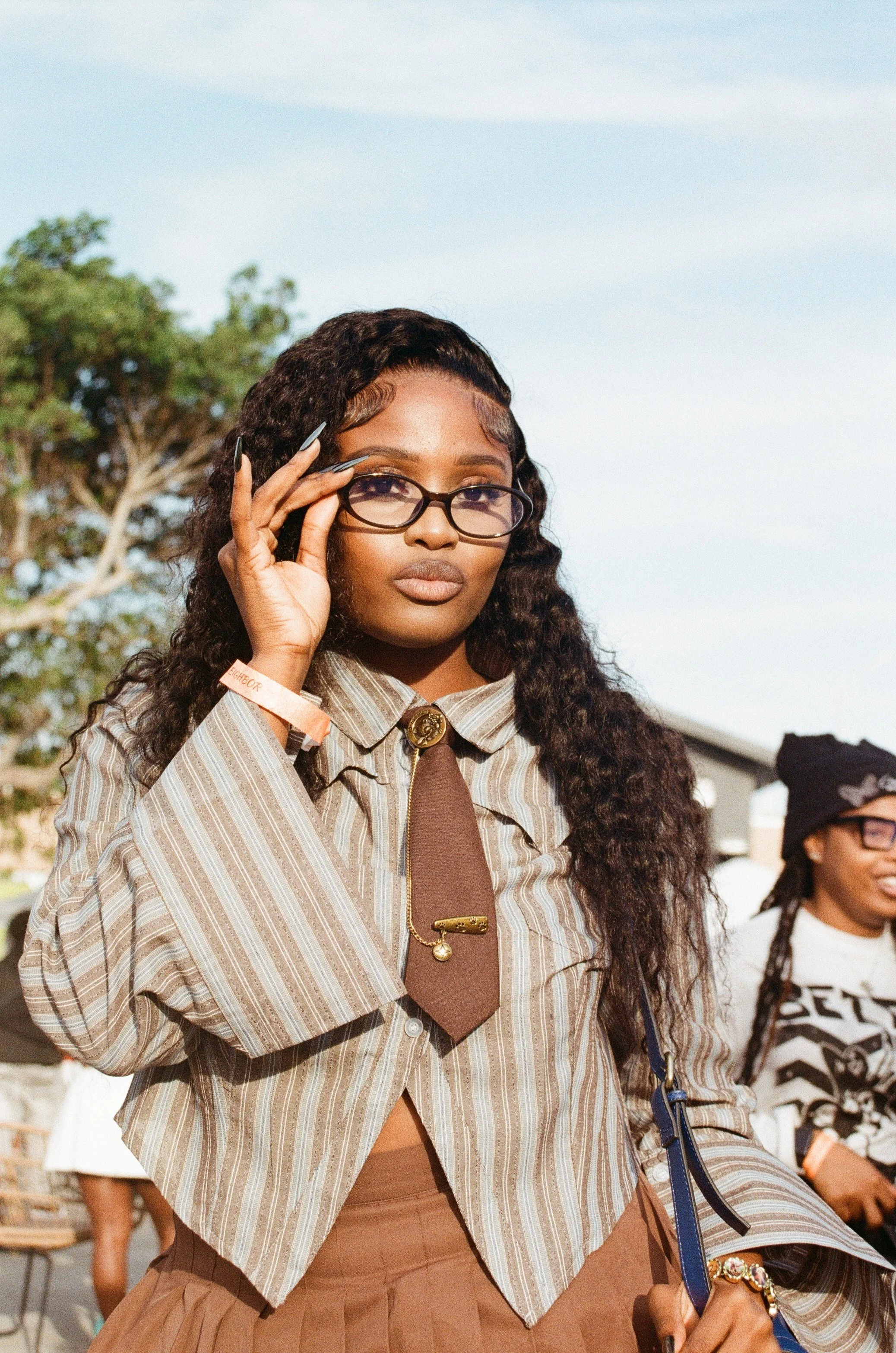 A woman adjusting her glasses outdoors with a clear sky and trees in the background. She has long, curly hair, wears a striped jacket, and a decorative tie with pins.