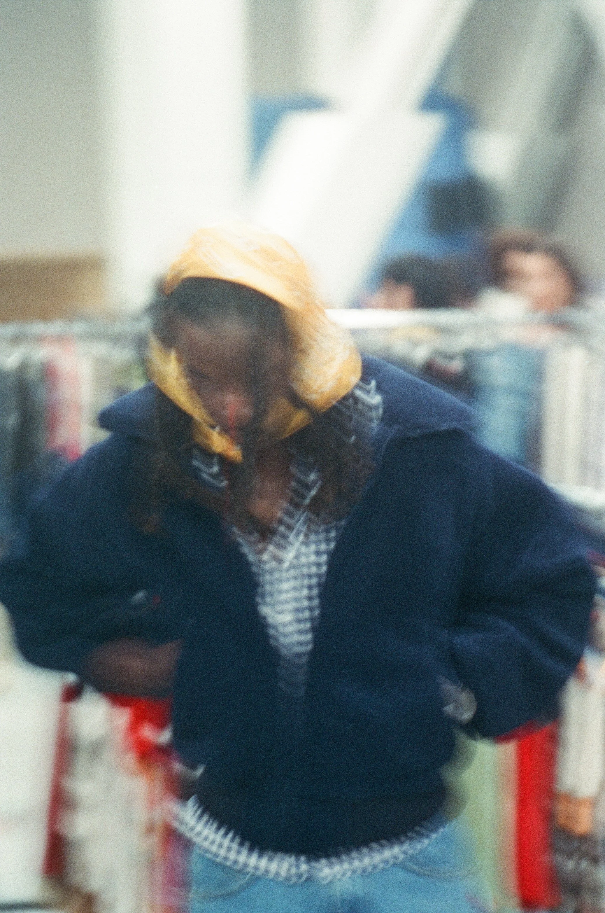 A woman with curly hair wearing a yellow hoodie under a navy jacket, looking down inside a bookstore or library.