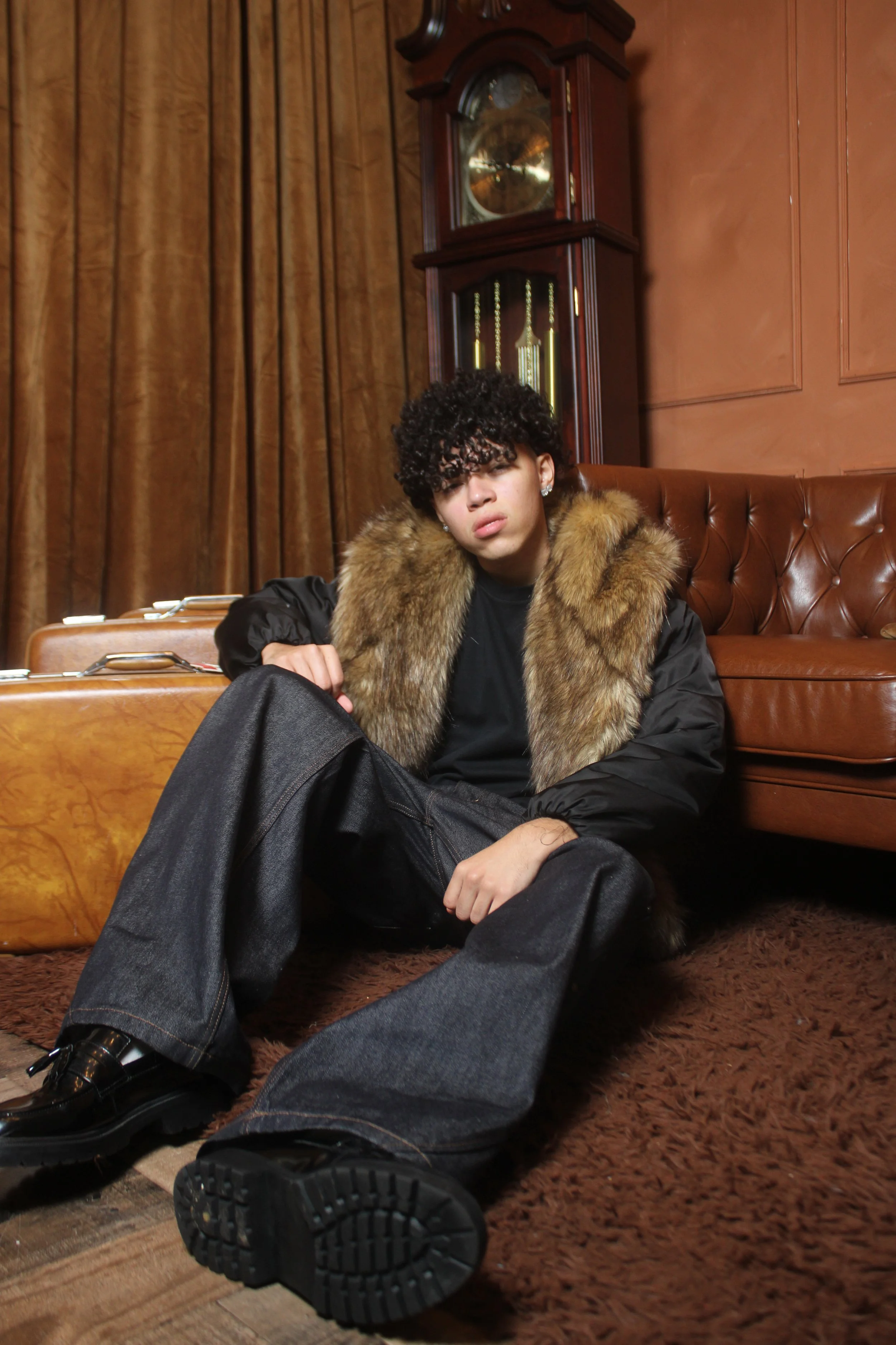Young man with curly hair sitting on the floor in a vintage living room, wearing a fur coat, black pants, and black shoes.