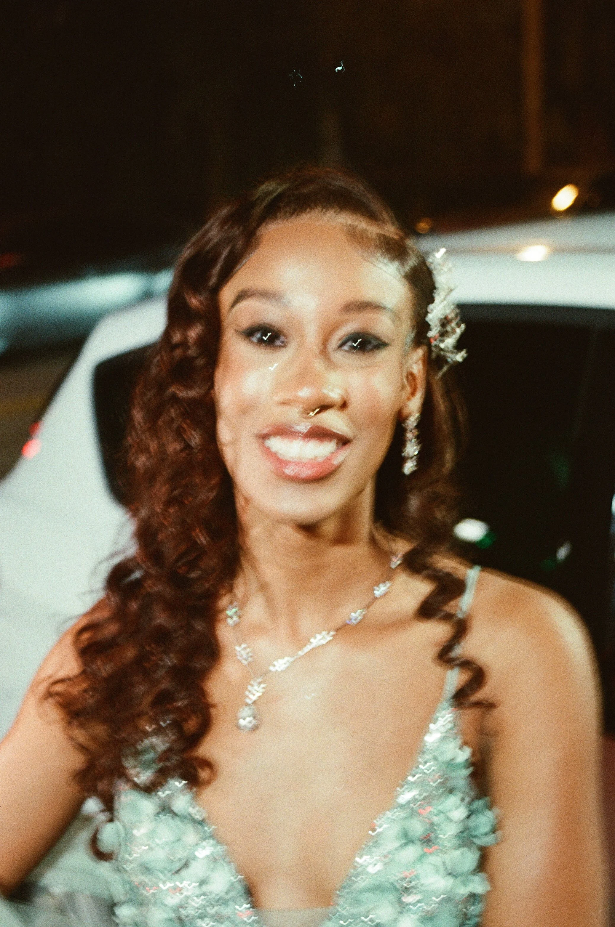 A young woman with long, curly brown hair smiling at the camera, dressed in a light-colored dress with floral embellishments and wearing jewelry.