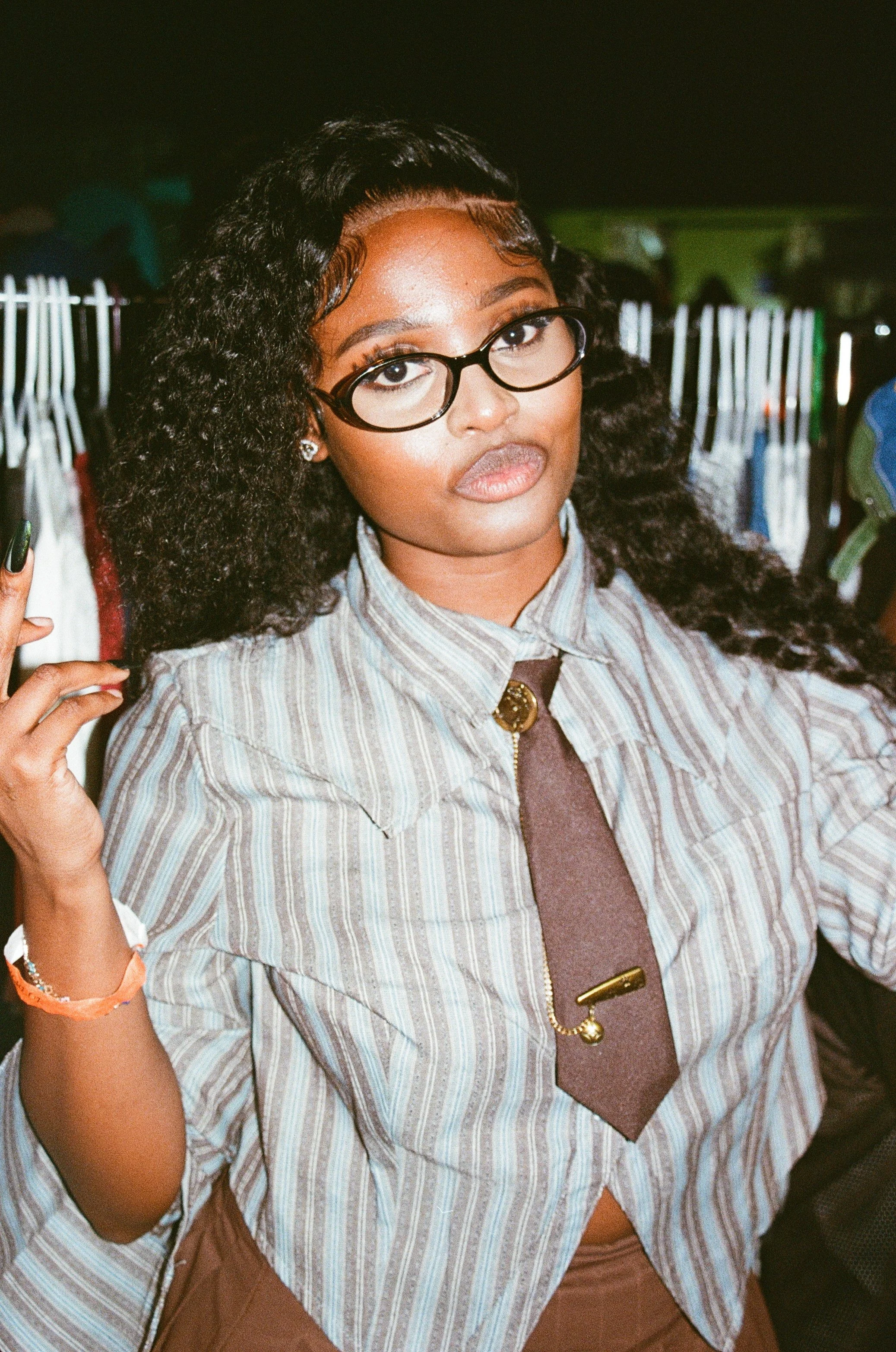 A young woman with curly black hair, glasses, and makeup, wearing a striped shirt and a brown tie with gold accents, standing in front of a clothing rack in a store or market.