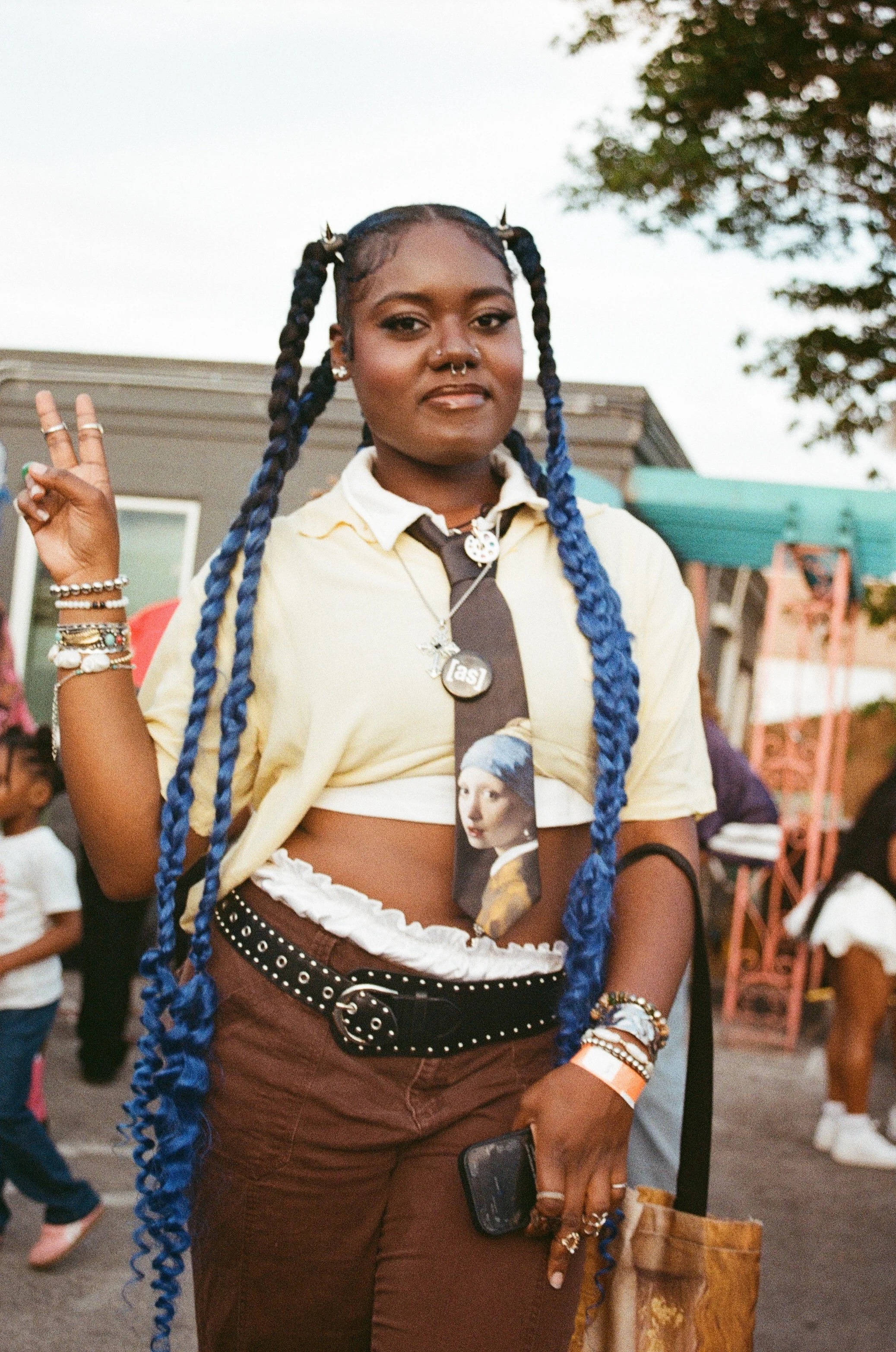 A young woman with long blue braided hair, wearing a light yellow crop top, a black tie with an image of a woman in a headscarf, and brown pants with eyelet details. She is accessorized with multiple rings, bracelets, and necklaces, and holding a pho