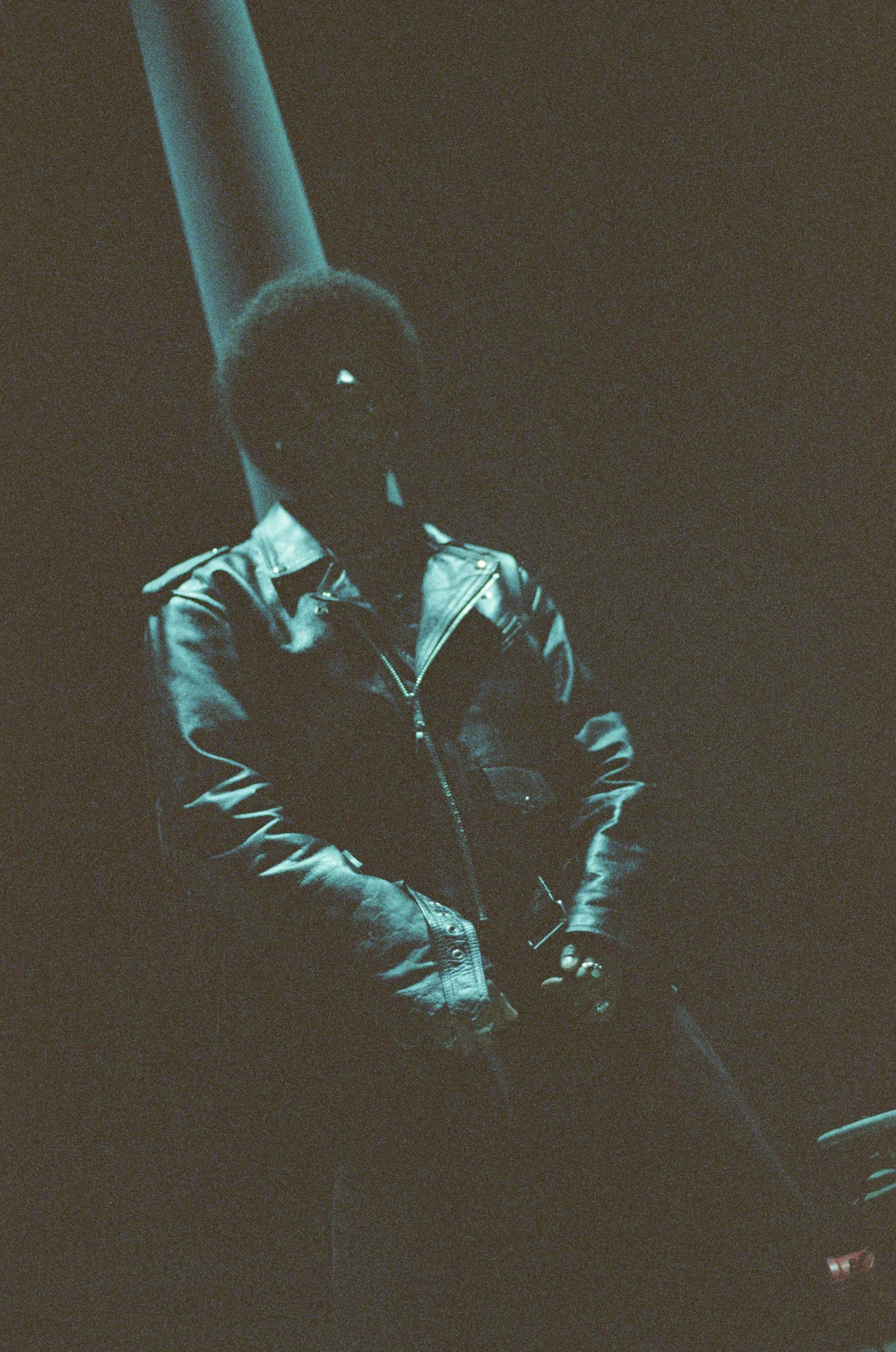 Person wearing a leather jacket and sunglasses, standing in a dark space with a spotlight shining from behind.