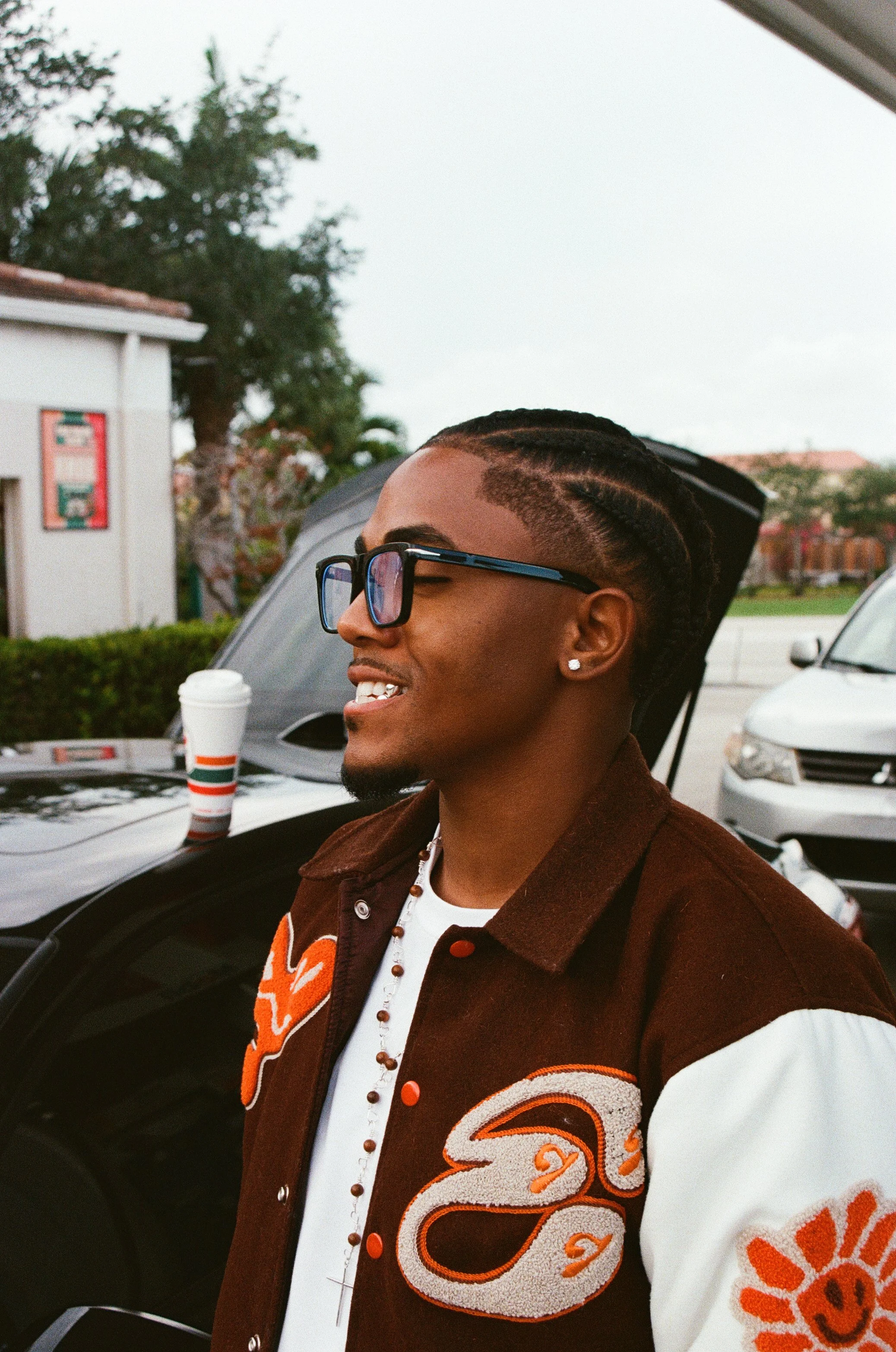 A young man with glasses and earrings smiling outdoors near parked cars, with a coffee cup on a car hood and trees in the background.