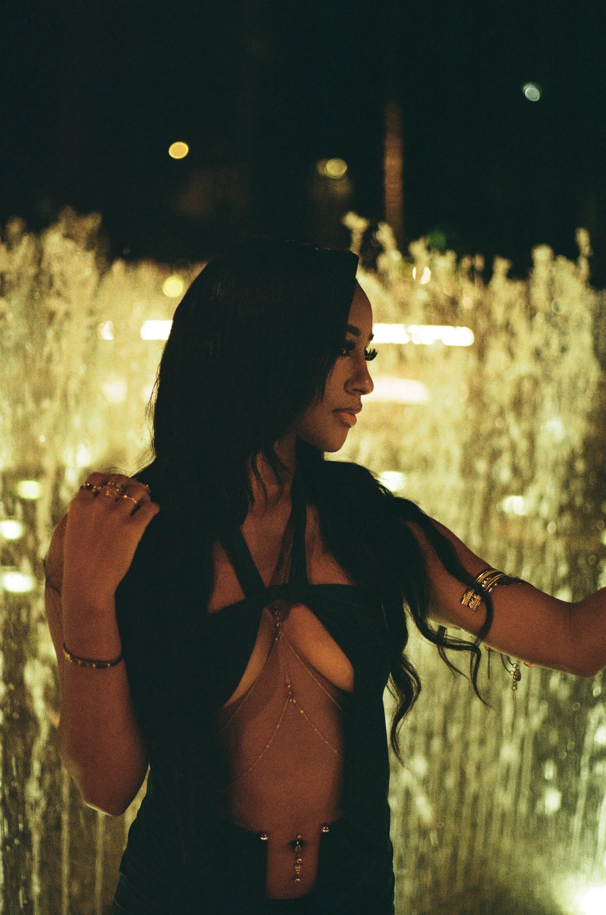 A woman with dark hair and jewelry standing in front of a lit fountain at night.