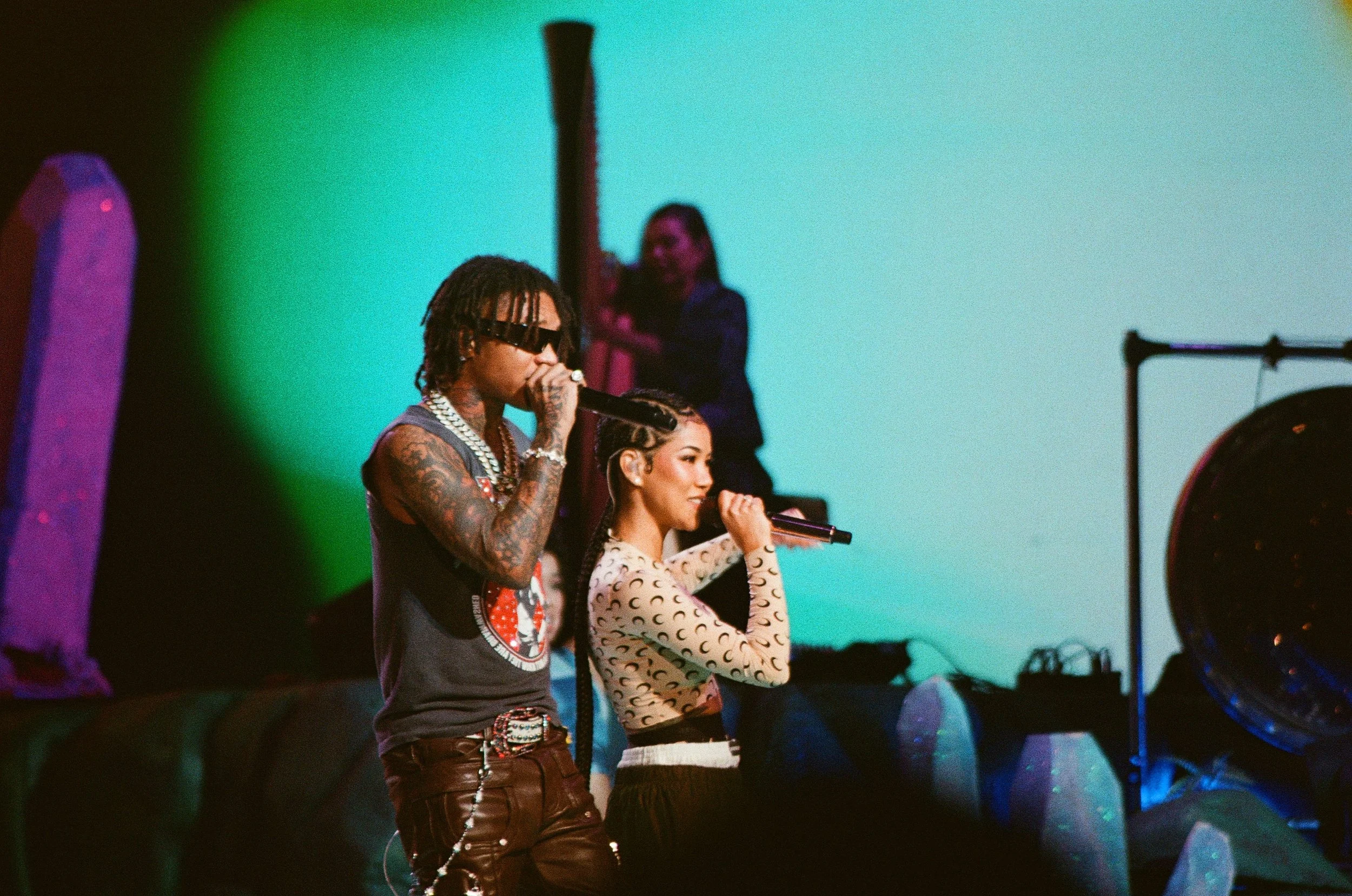 Two performers, a man and a woman, singing into microphones on stage with colorful lighting, while a woman in the background holds a tall object.