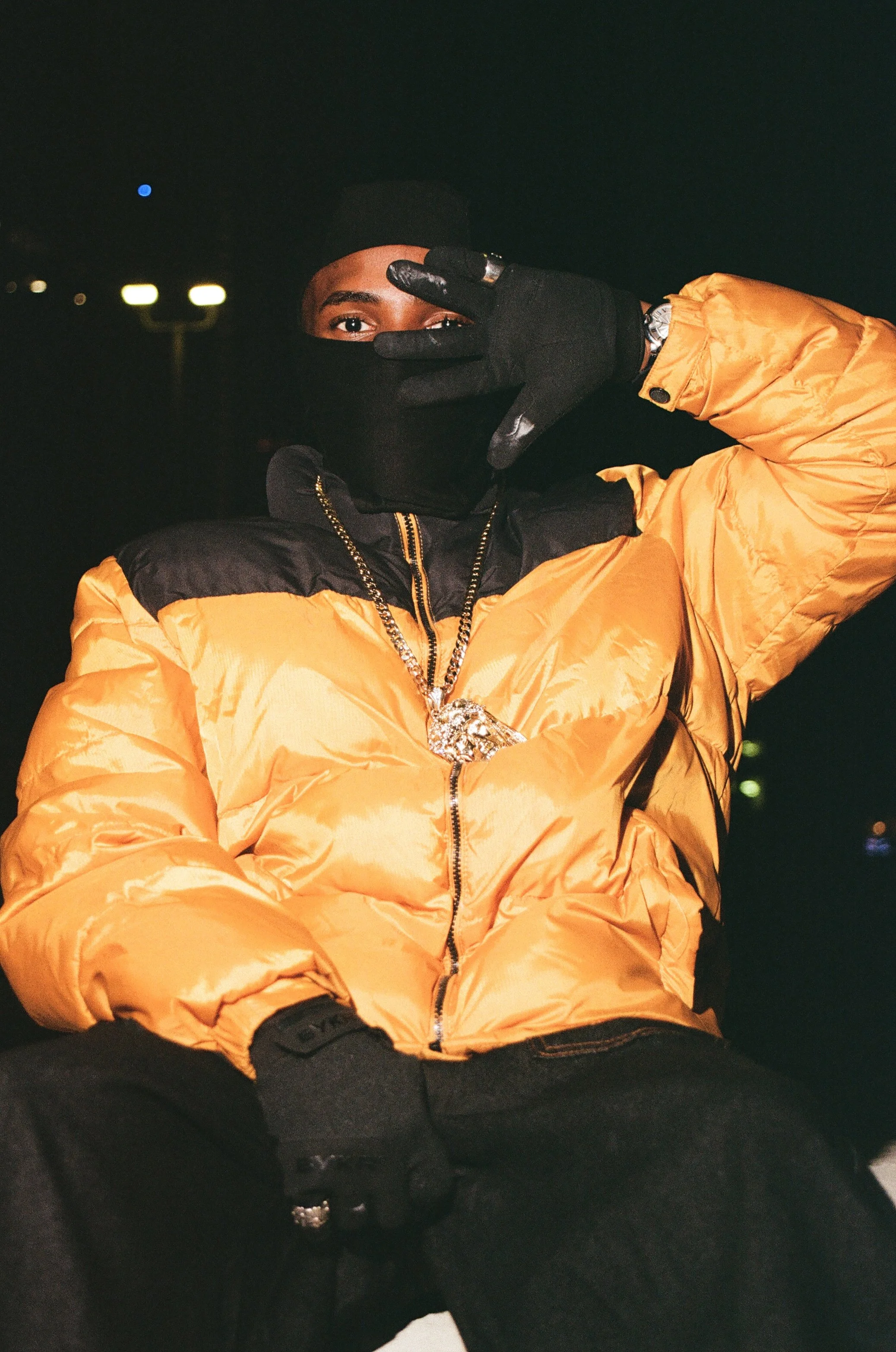 A person wearing a black face mask, black gloves, a yellow puffer jacket with black accents, and a large chain necklace, posing with one hand over their face at night.