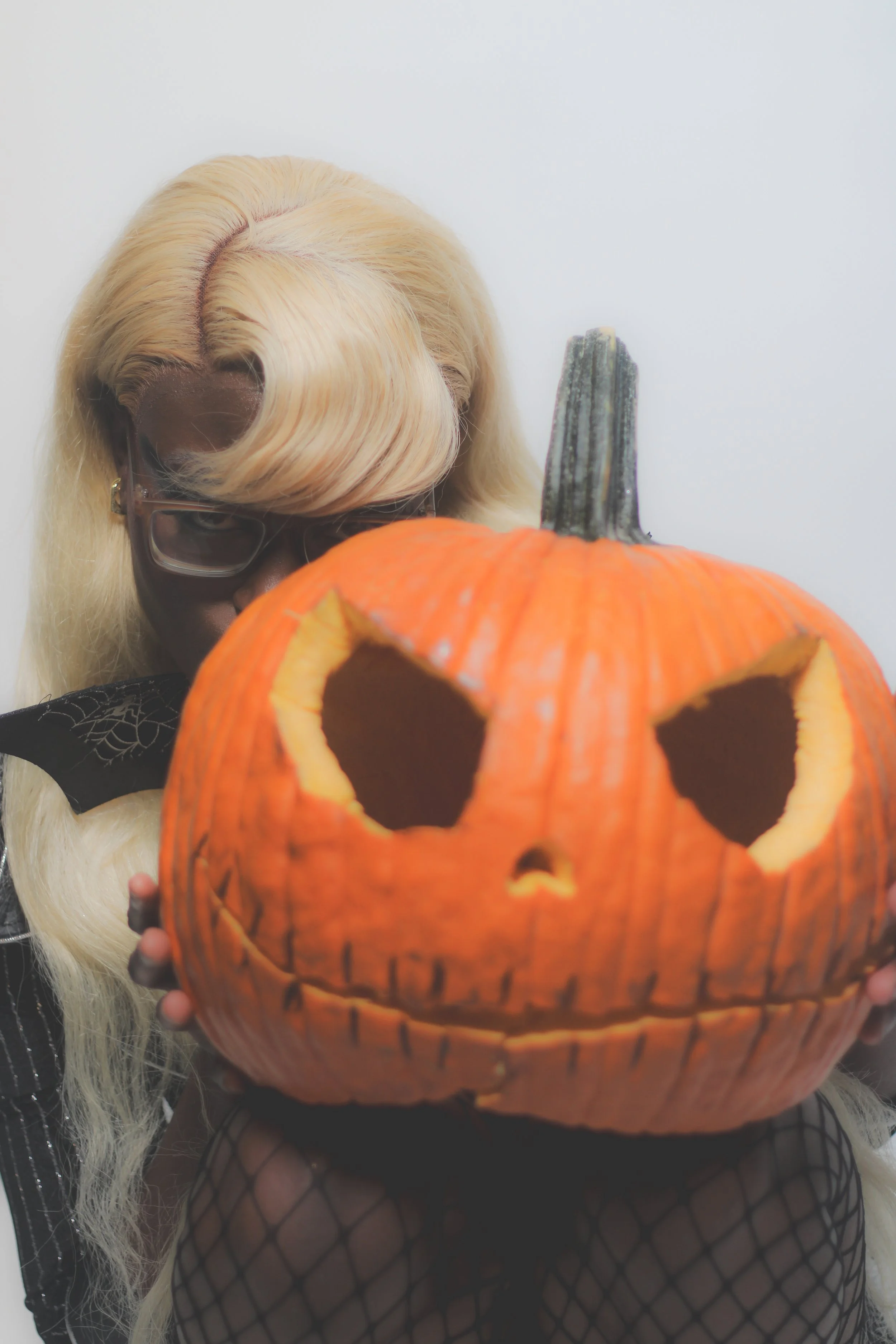 Person with blonde hair, glasses, wearing black fishnet gloves, holding a carved pumpkin with triangular eyes and a stitched mouth, facing the camera.