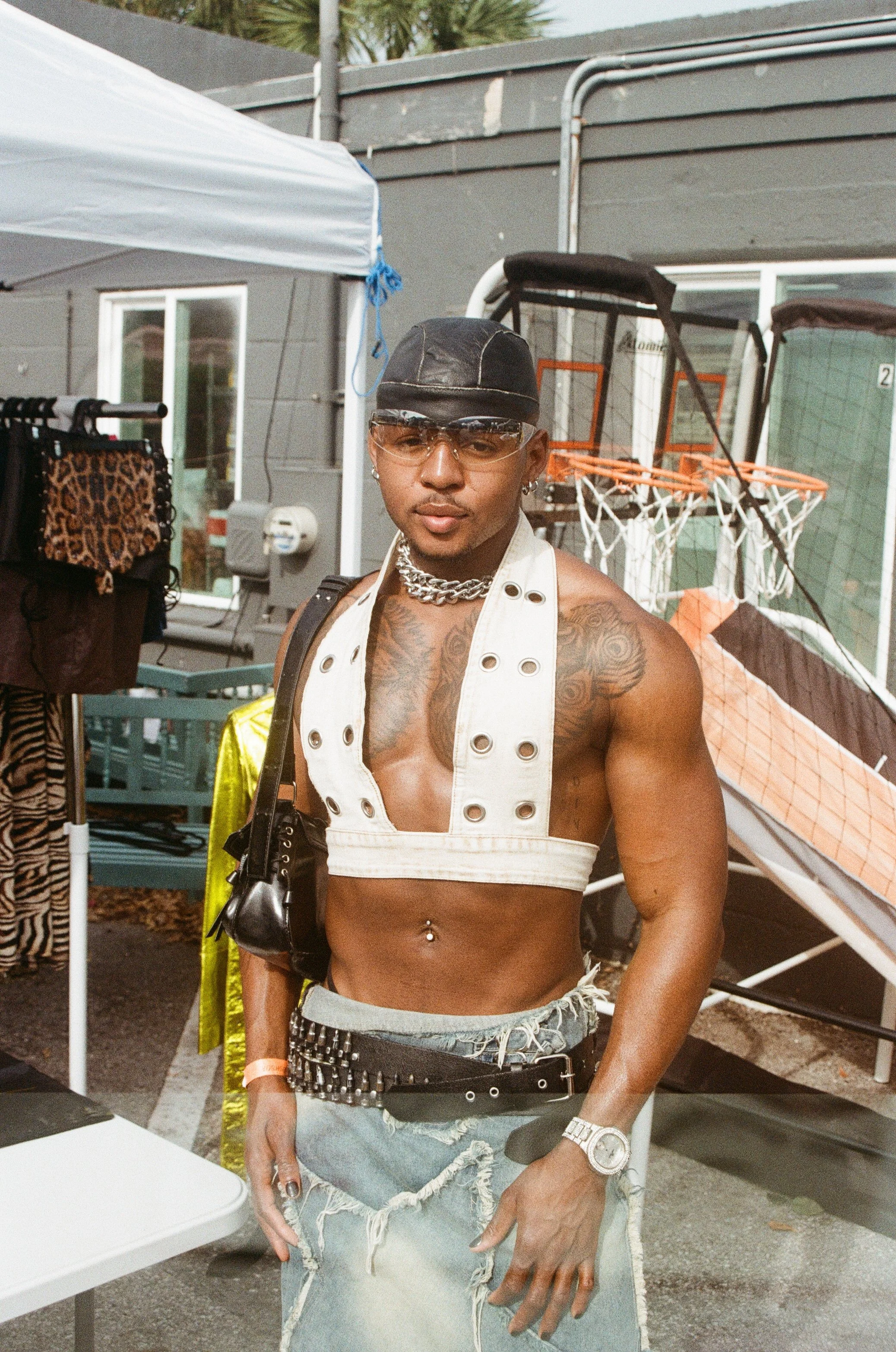A shirtless man with tattoos, wearing a white crop top, distressed denim shorts, a black cap, sunglasses, and jewelry, standing outdoors near a clothing rack and a basketball hoop.