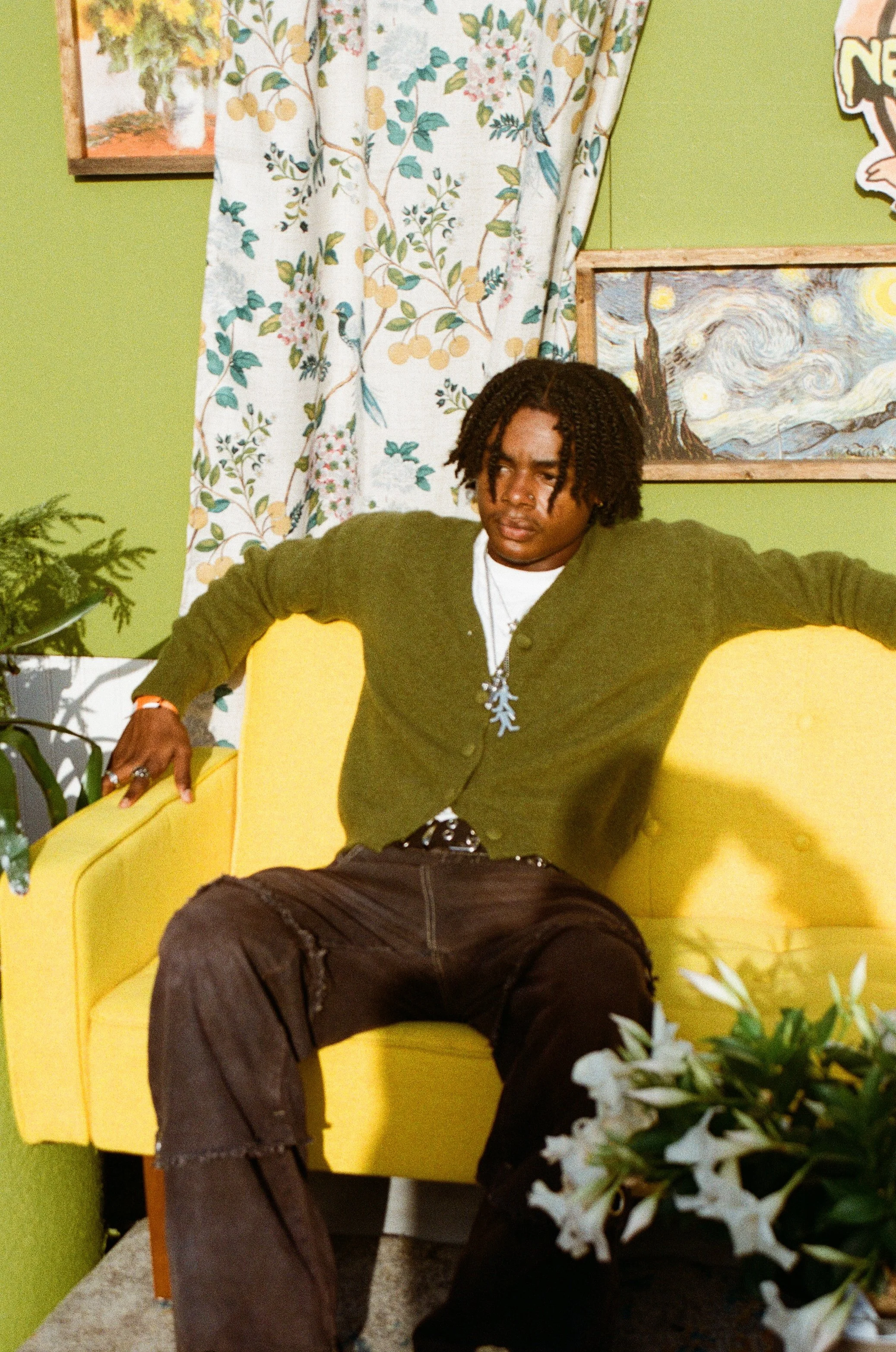 A young man with dreadlocks sitting on a bright yellow armchair in a room with green walls, floral curtains, and framed artwork including a Van Gogh painting.