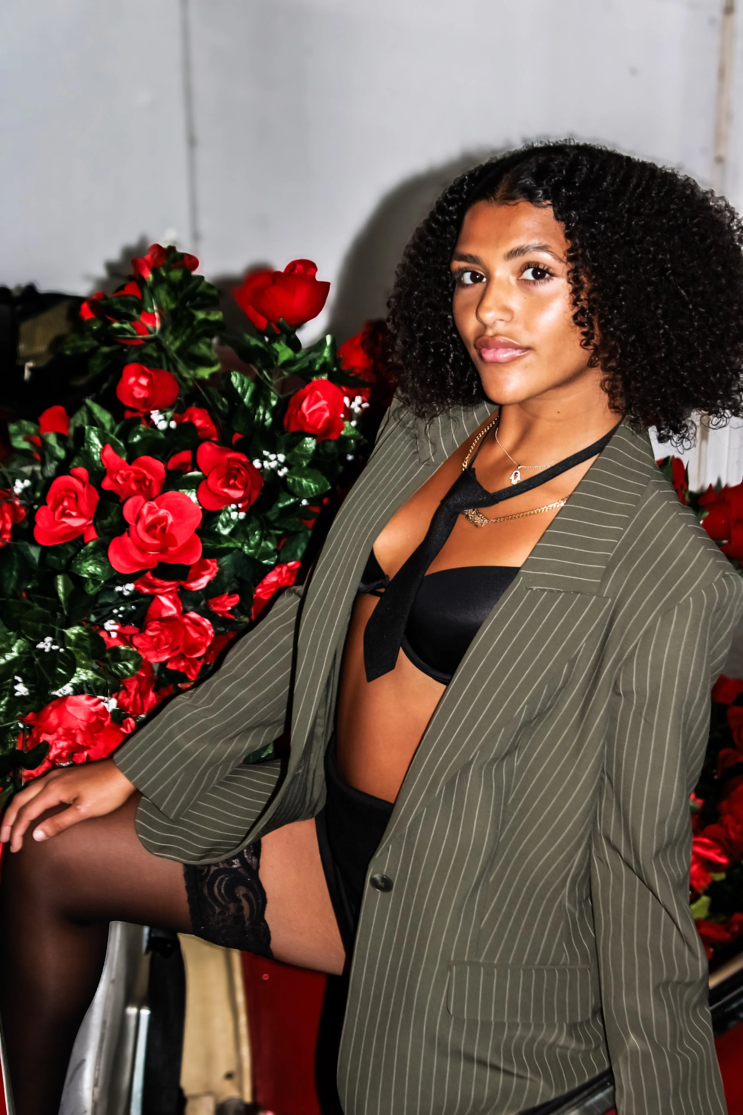 A woman with curly hair wearing a pinstriped blazer, black lingerie, and black thigh-high stockings, sitting on a wheelchair near a large bouquet of red roses.