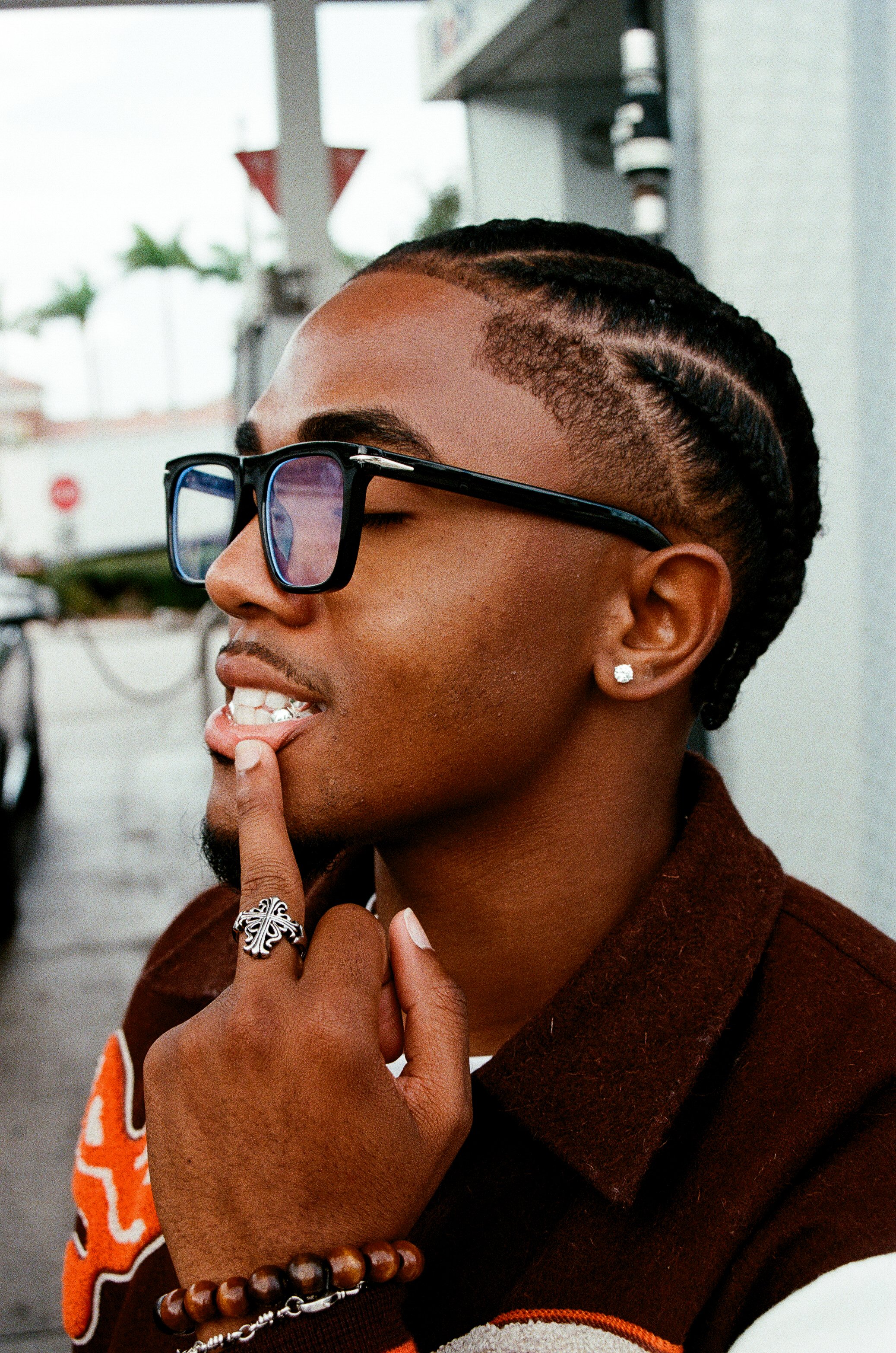 Close-up of a young man with braided hair and sunglasses, touching his lips with his finger, wearing a brown jacket with orange accents and jewelry.