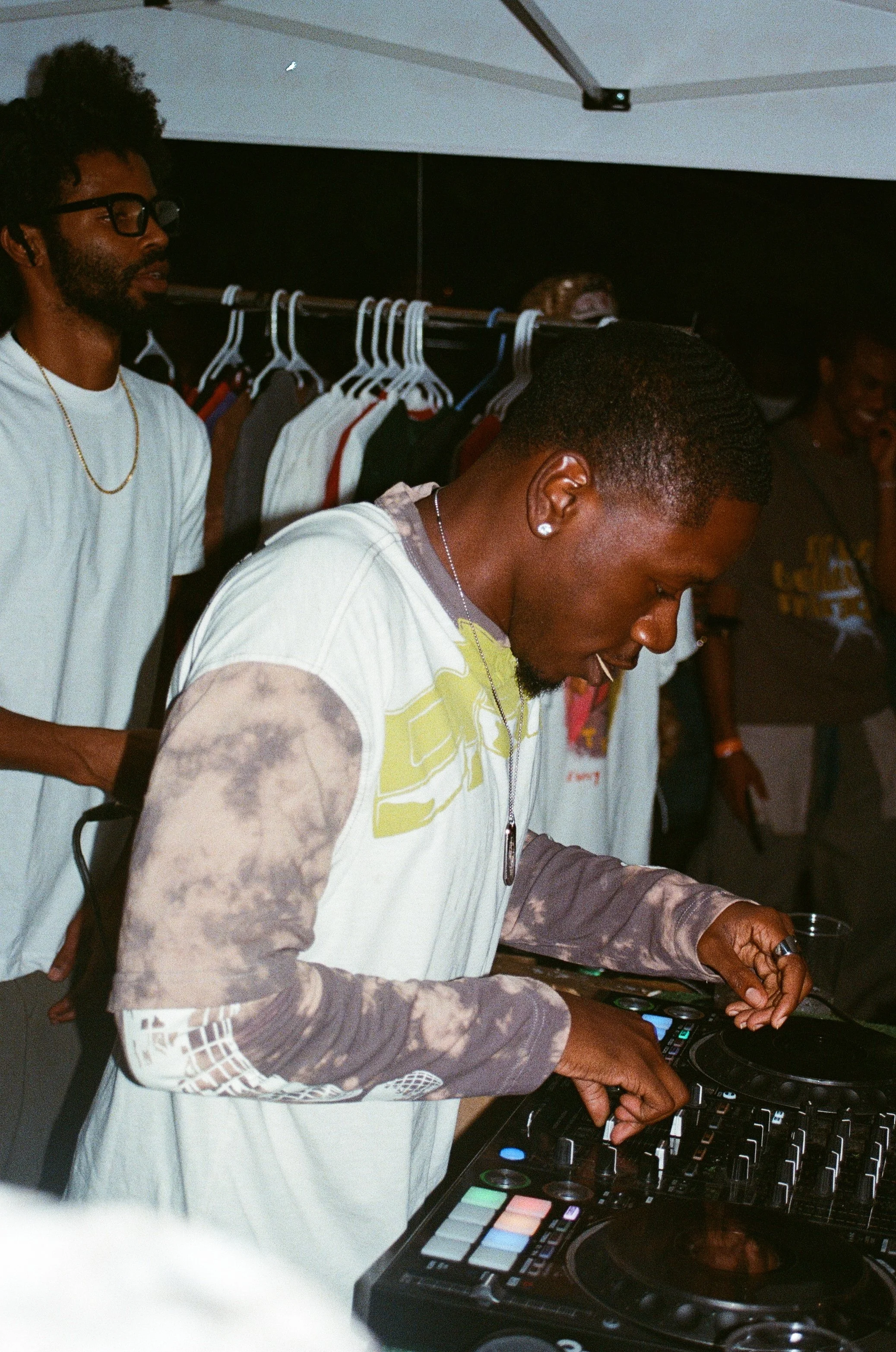 A young male DJ with short hair and a beard, wearing a white and gray tie-dye long-sleeve shirt, is mixing music on a DJ controller at an outdoor party at night. There are other people in the background, some looking at him and others talking.