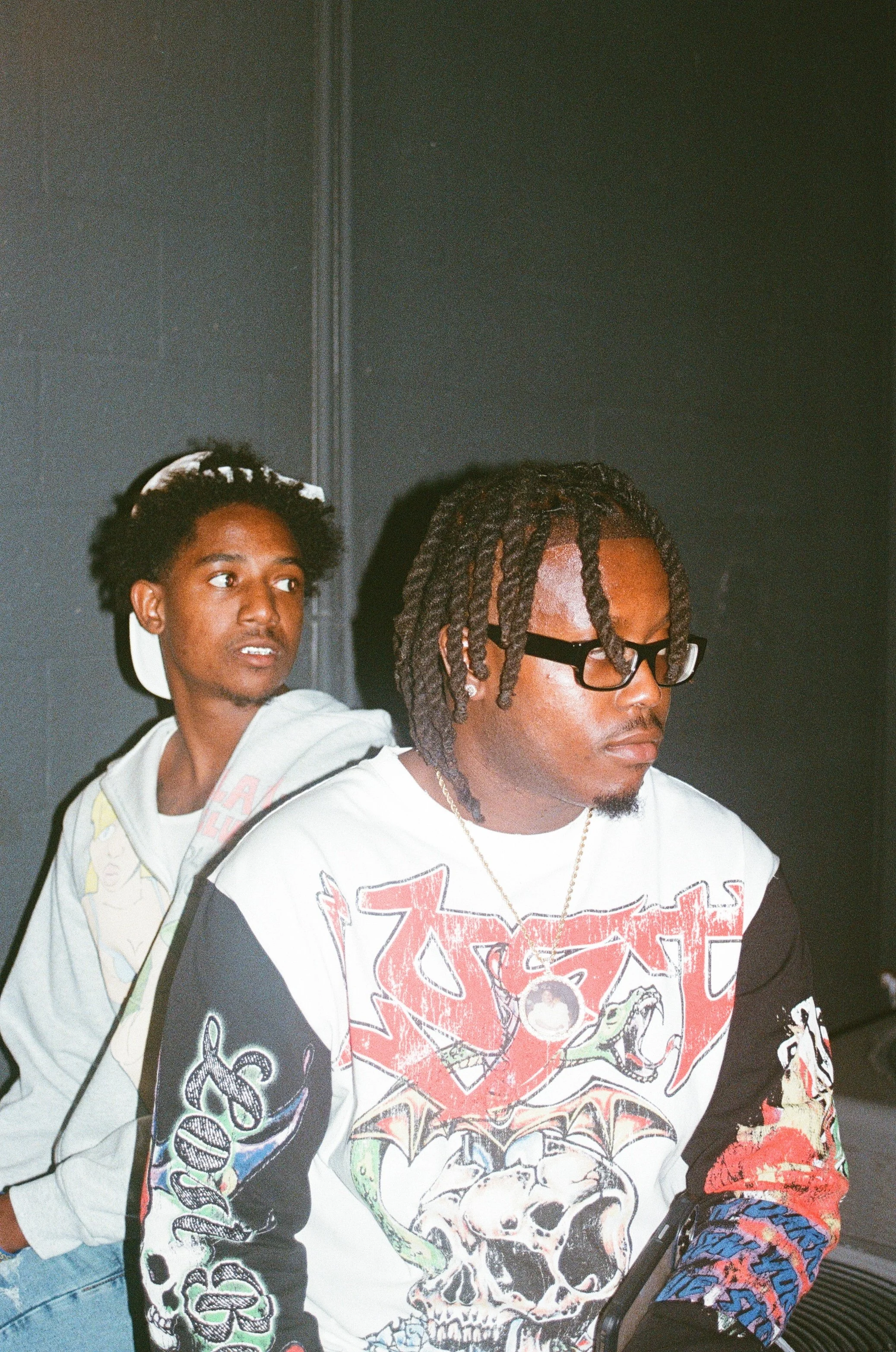 Two young men sitting against a dark wall, one with braided hair and glasses, wearing a graphic shirt featuring skulls and dragons, and the other with curly hair, a headband, and a light hoodie.
