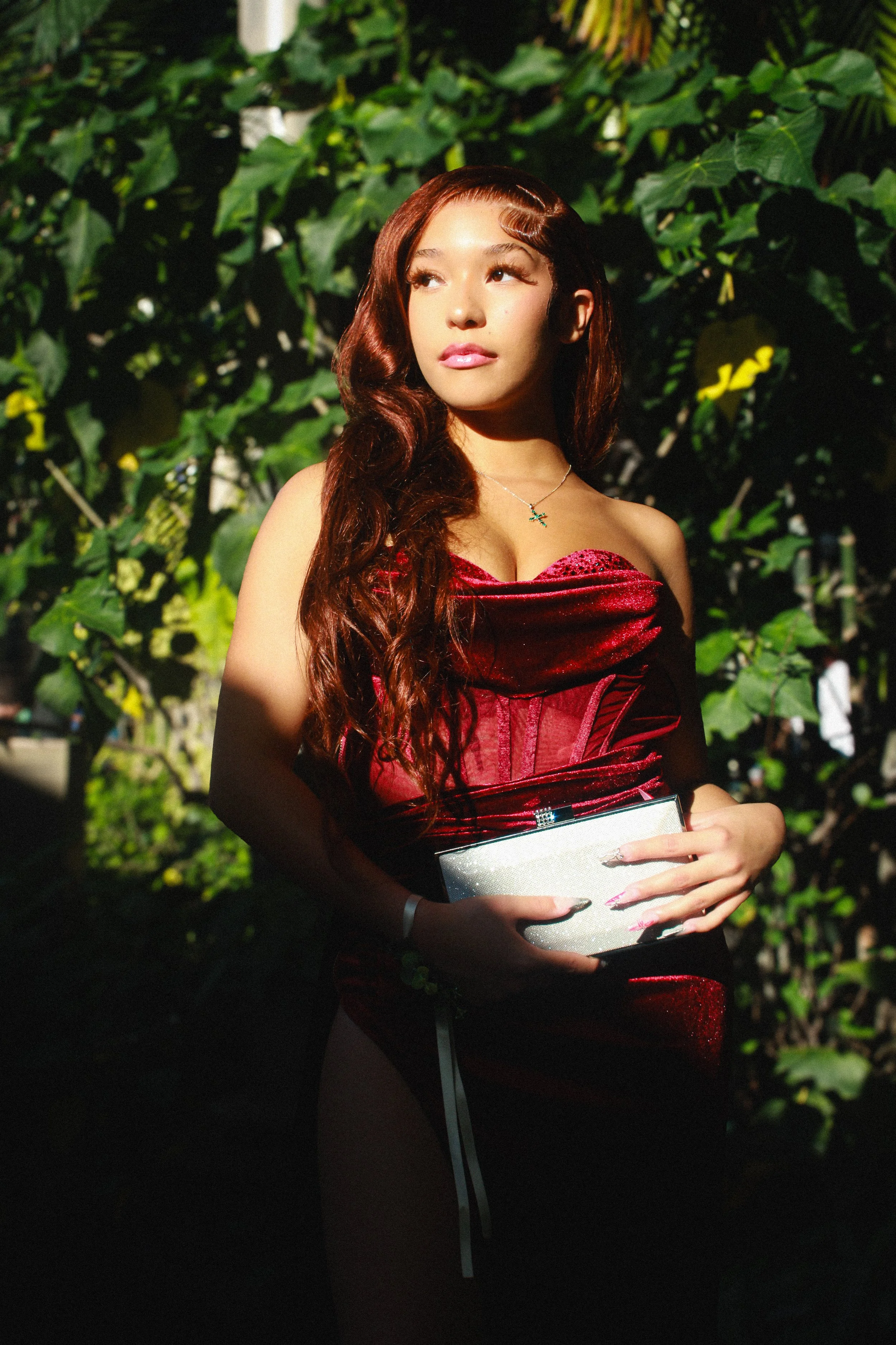 A woman with long, wavy brown hair holding a silver clutch, standing in a lush, green outdoor setting, wearing a strapless red dress.