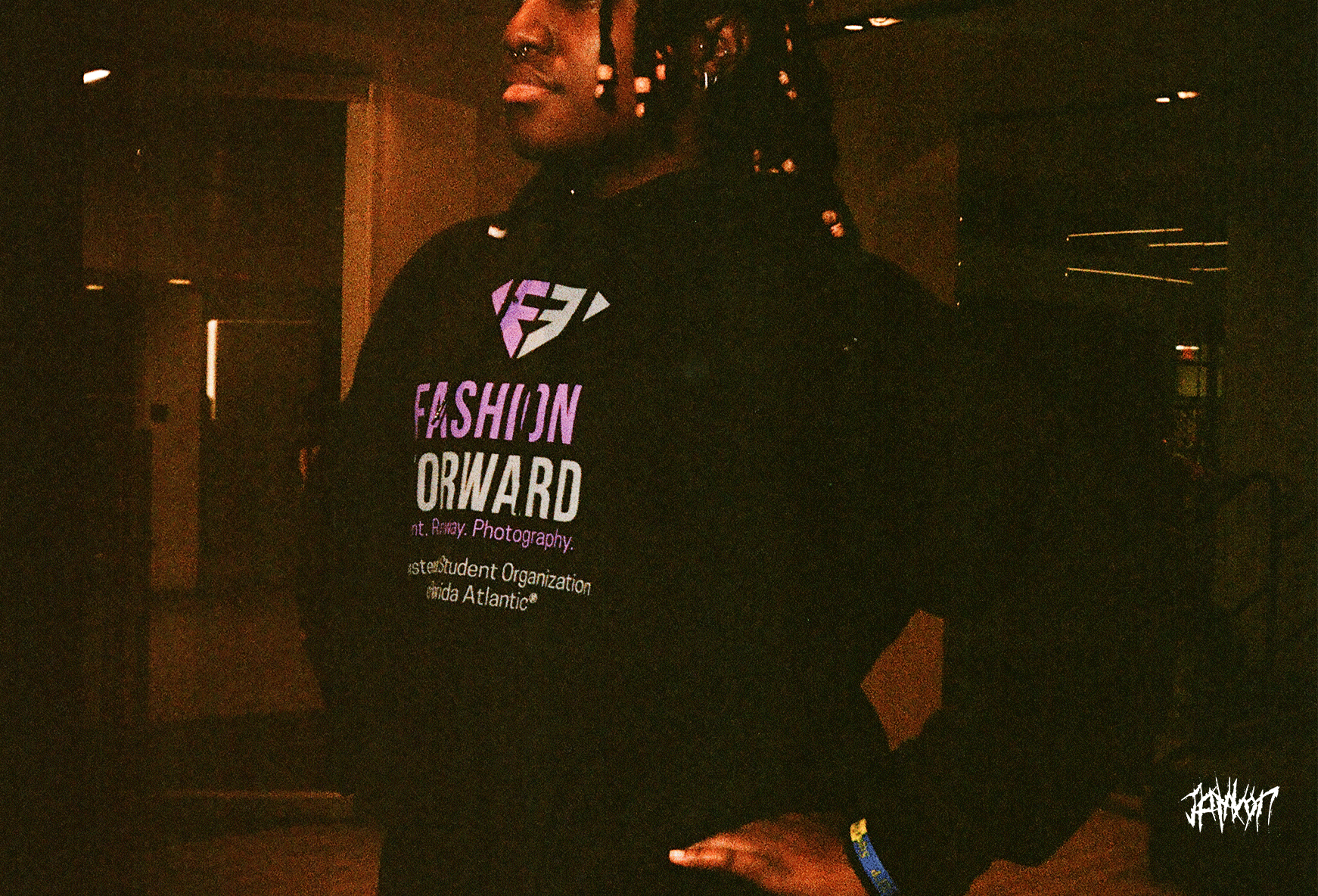 Person wearing a black hoodie with colorful text and graphics, dark indoor setting, photographer's signature in the bottom right corner.