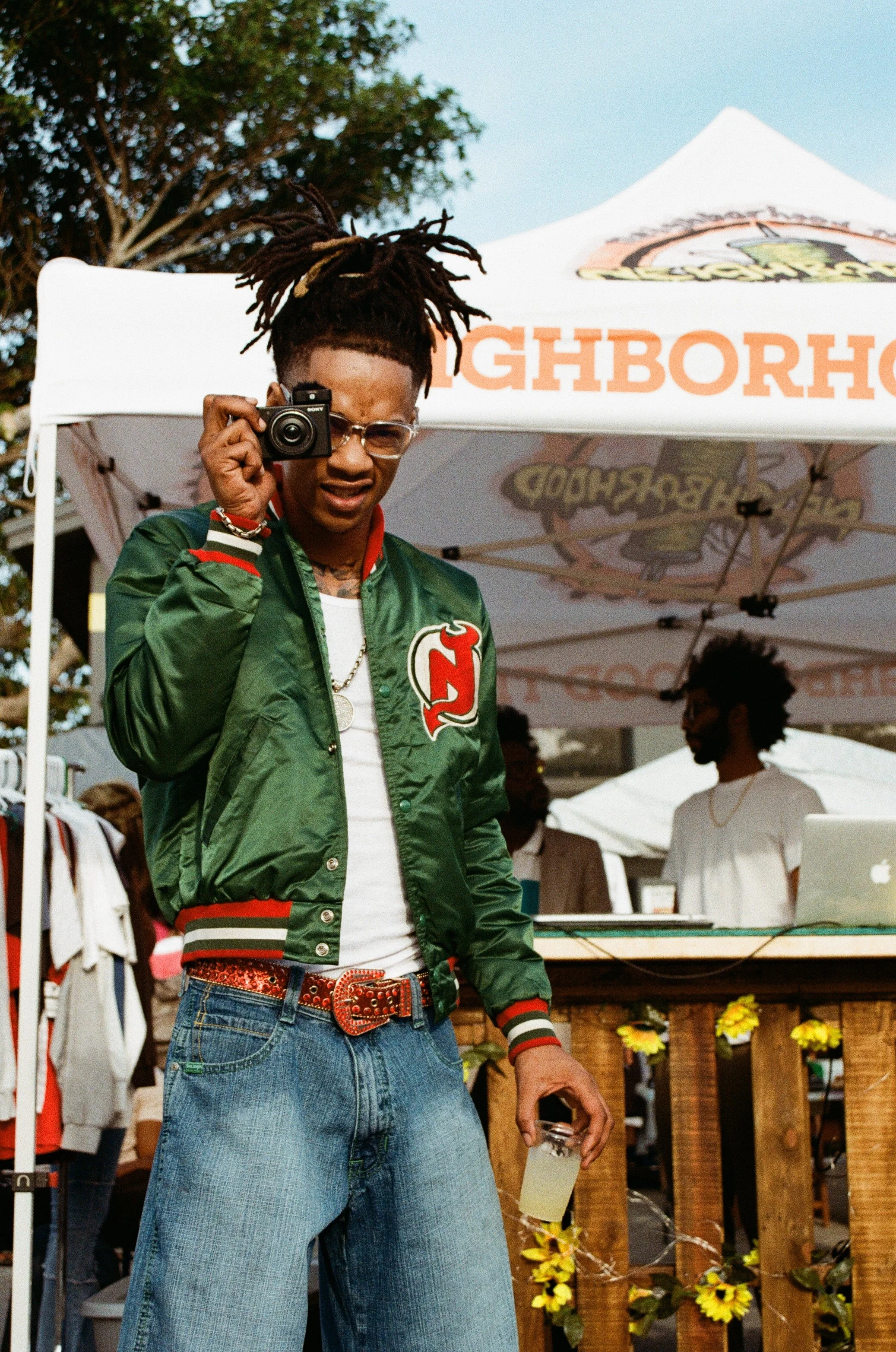 A young man with dreadlocks, glasses, and a green jacket, taking a photo with a camera, standing outdoors at a lively event with tents and people in the background.