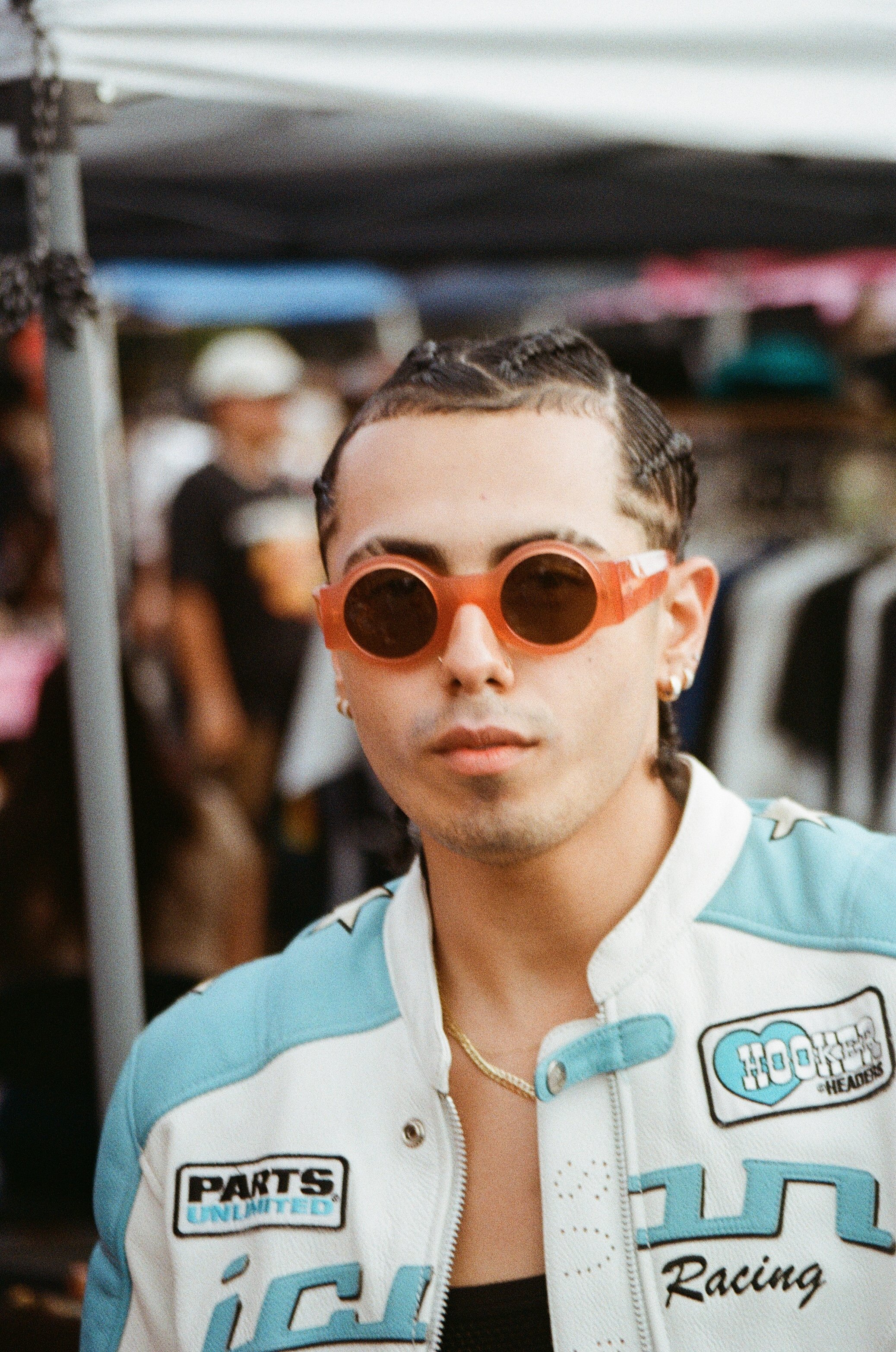 A young person with braided hair wearing red round sunglasses, earrings, a necklace, and a racing jacket with patches, outdoors at a populated event.