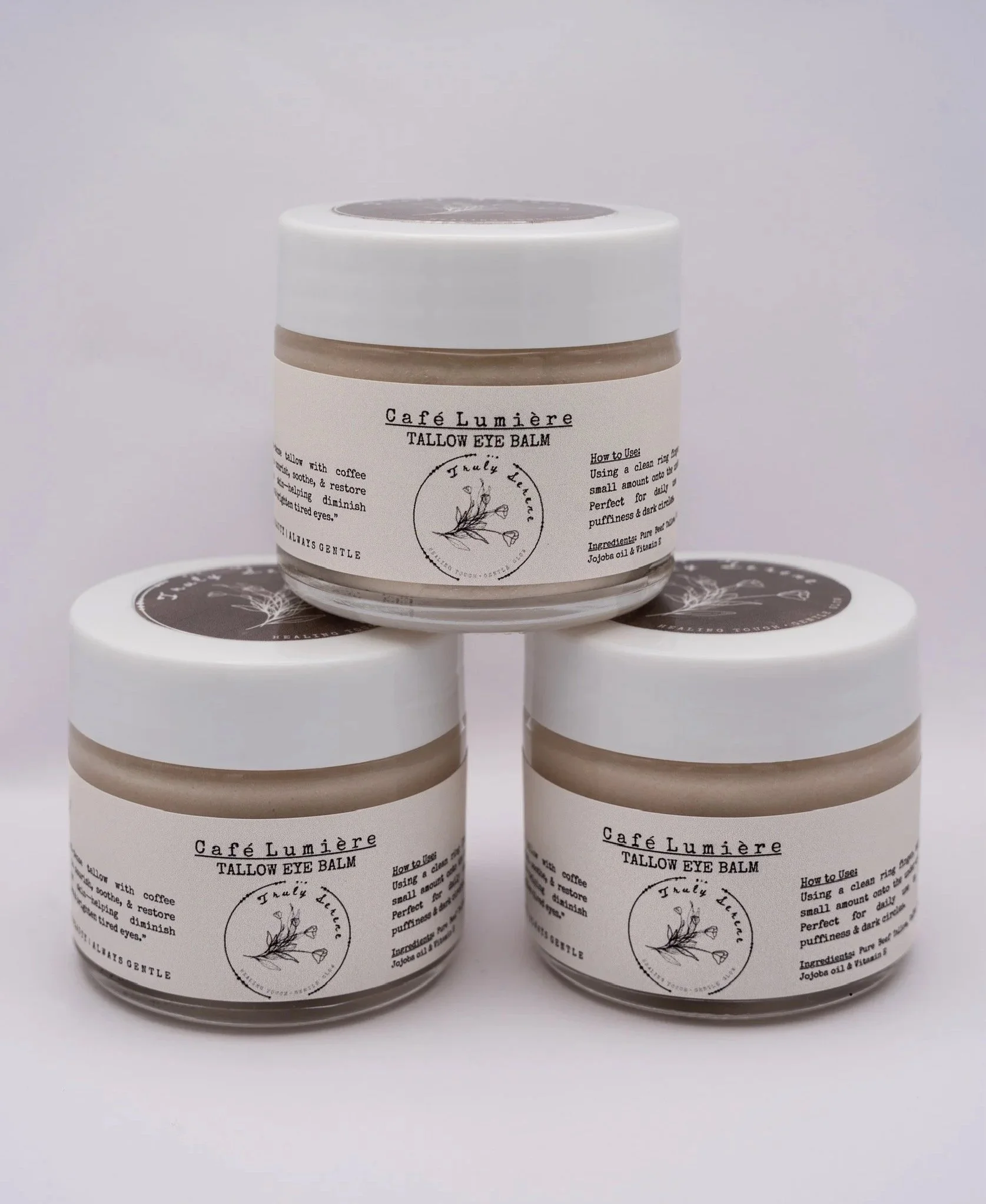 Three jars of Café Lumière Tallow Eye Balm stacked in a pyramid shape against a plain background.