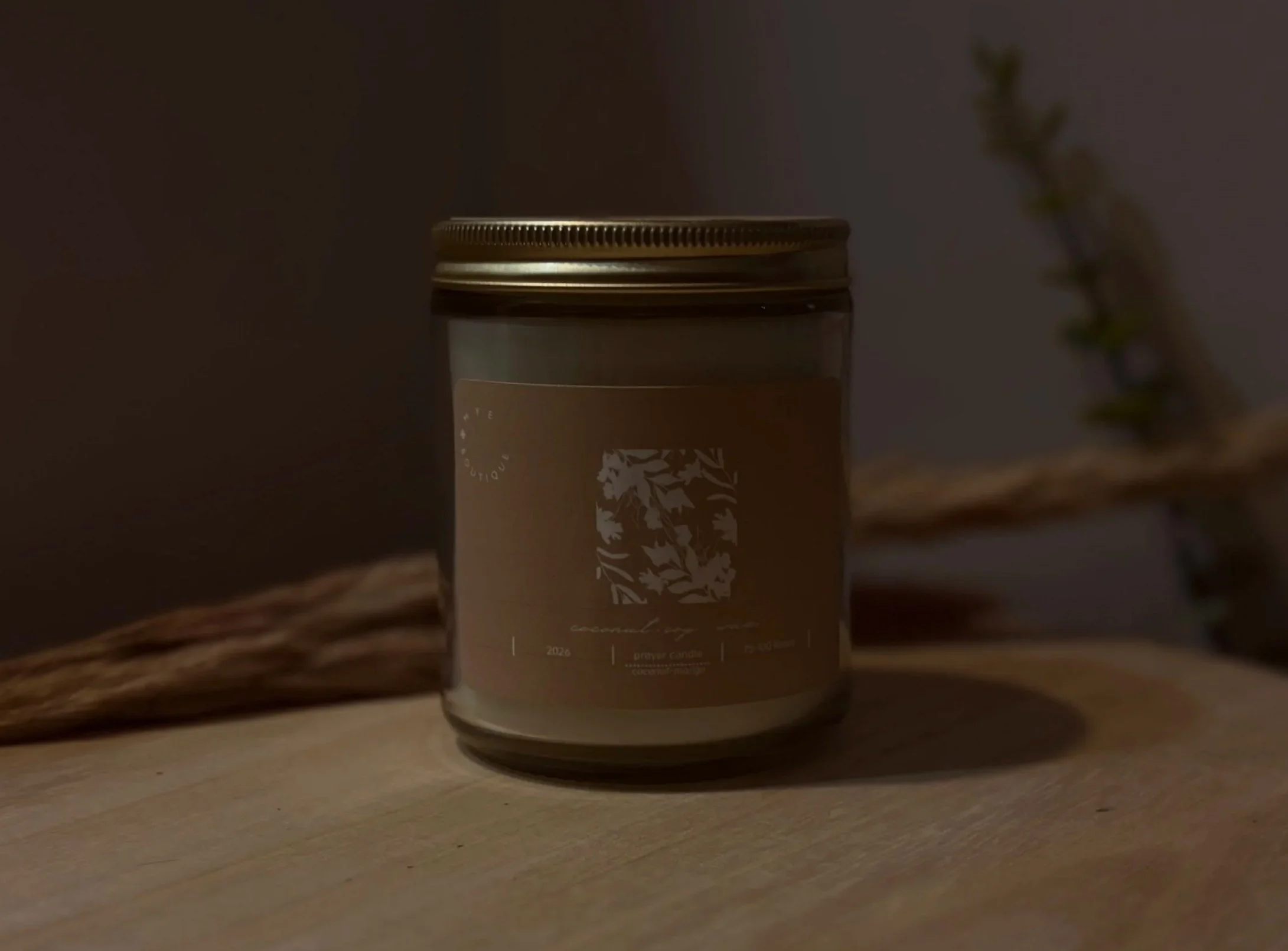 A candle jar with a metallic lid placed on a wooden surface, with a blurred plant in the background.