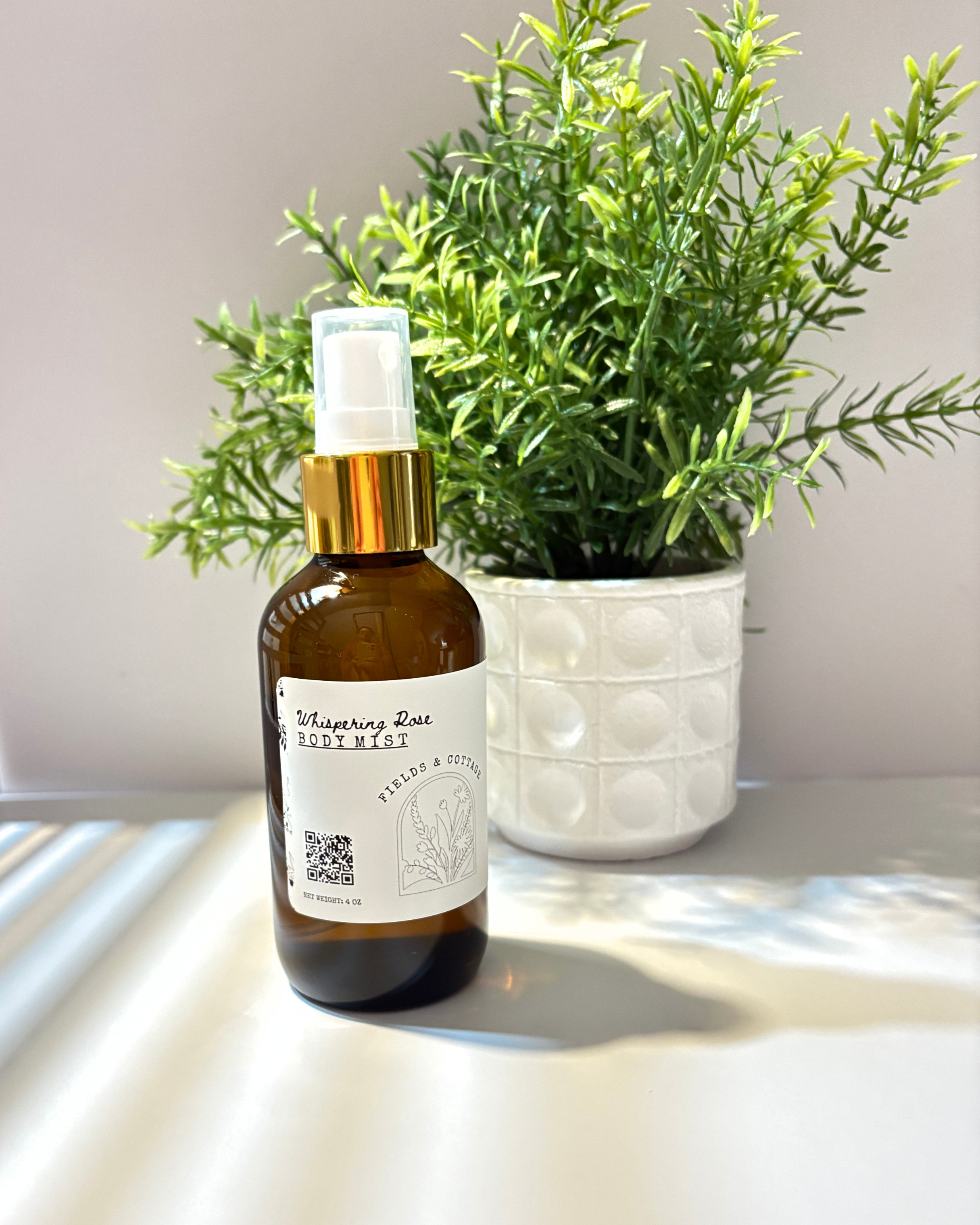A brown glass spray bottle labeled 'Whispering Rose Body Mist' with a gold spray top, placed on a white surface in front of a potted green plant with dense, elongated leaves.