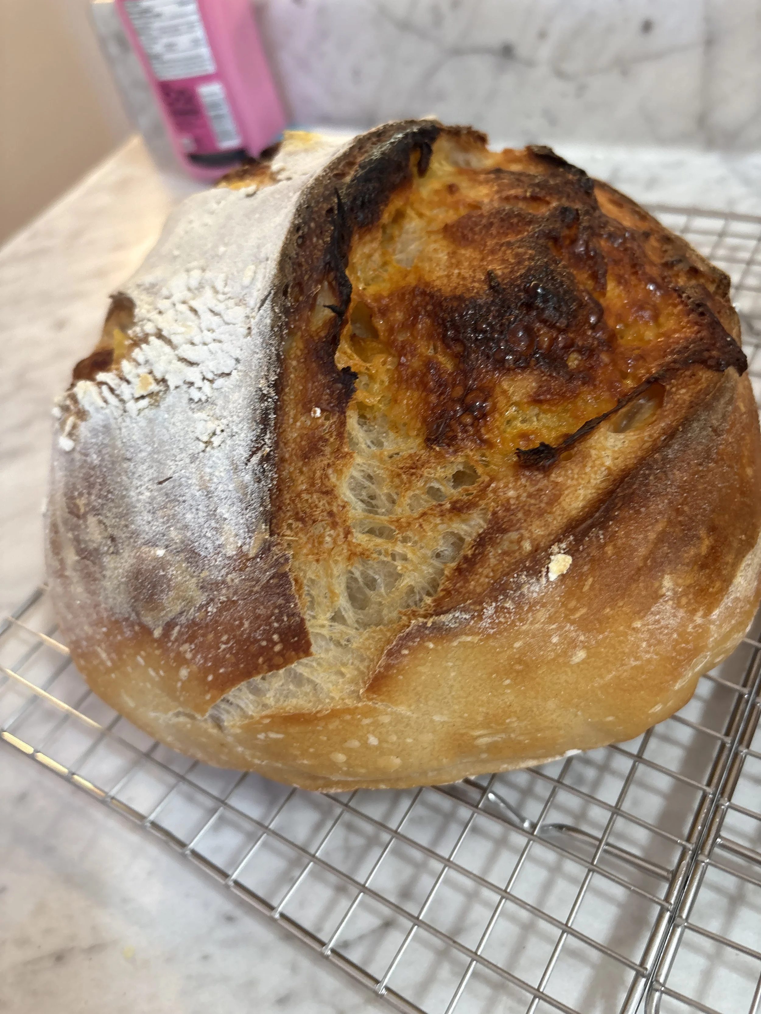 A close-up of a freshly baked loaf of bread with a golden-brown crust, resting on a cooling rack, with part of a pink beverage or packaging in the background.