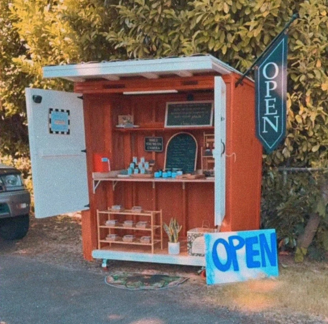 A small wooden stand with a sign that says 'OPEN' and a tall hanging sign also saying 'OPEN'. Inside the stand, there are shelves with various small items, a chalkboard, and a small potted plant outside. There is a vehicle partially visible to the left, and trees in the background.