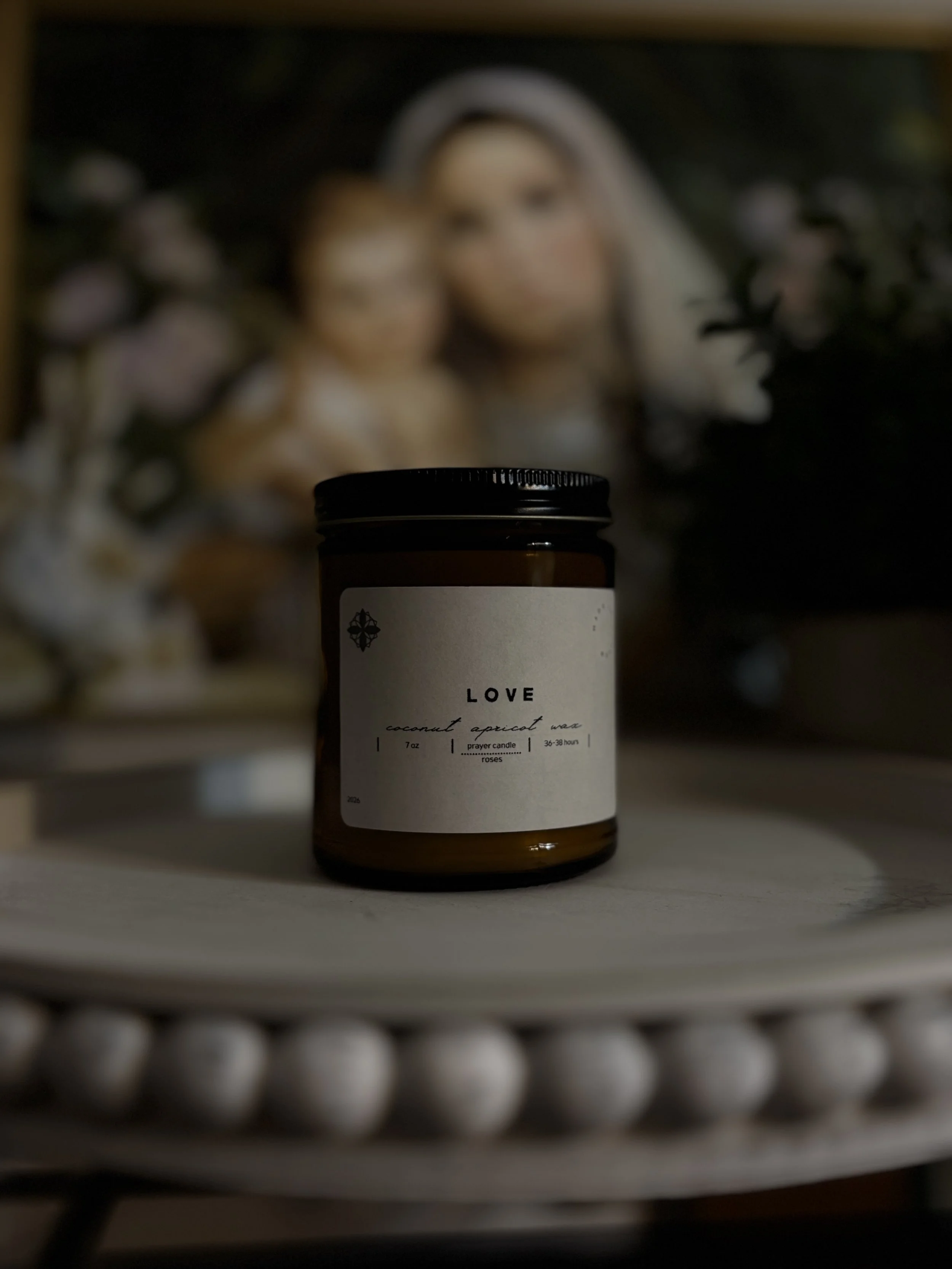A brown candle jar labeled 'LOVE' sitting on a white round table with a blurred background showing a portrait of a woman and a child.
