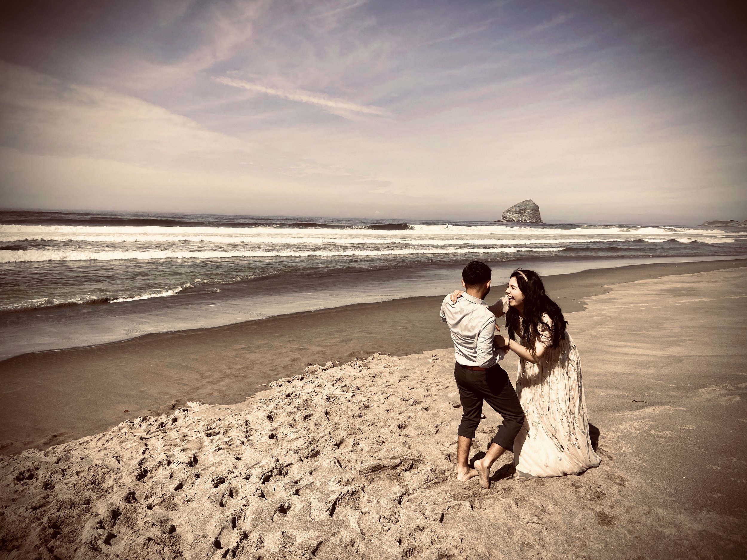 A couple dancing on a beach with ocean waves and a large rock formation in the distance.