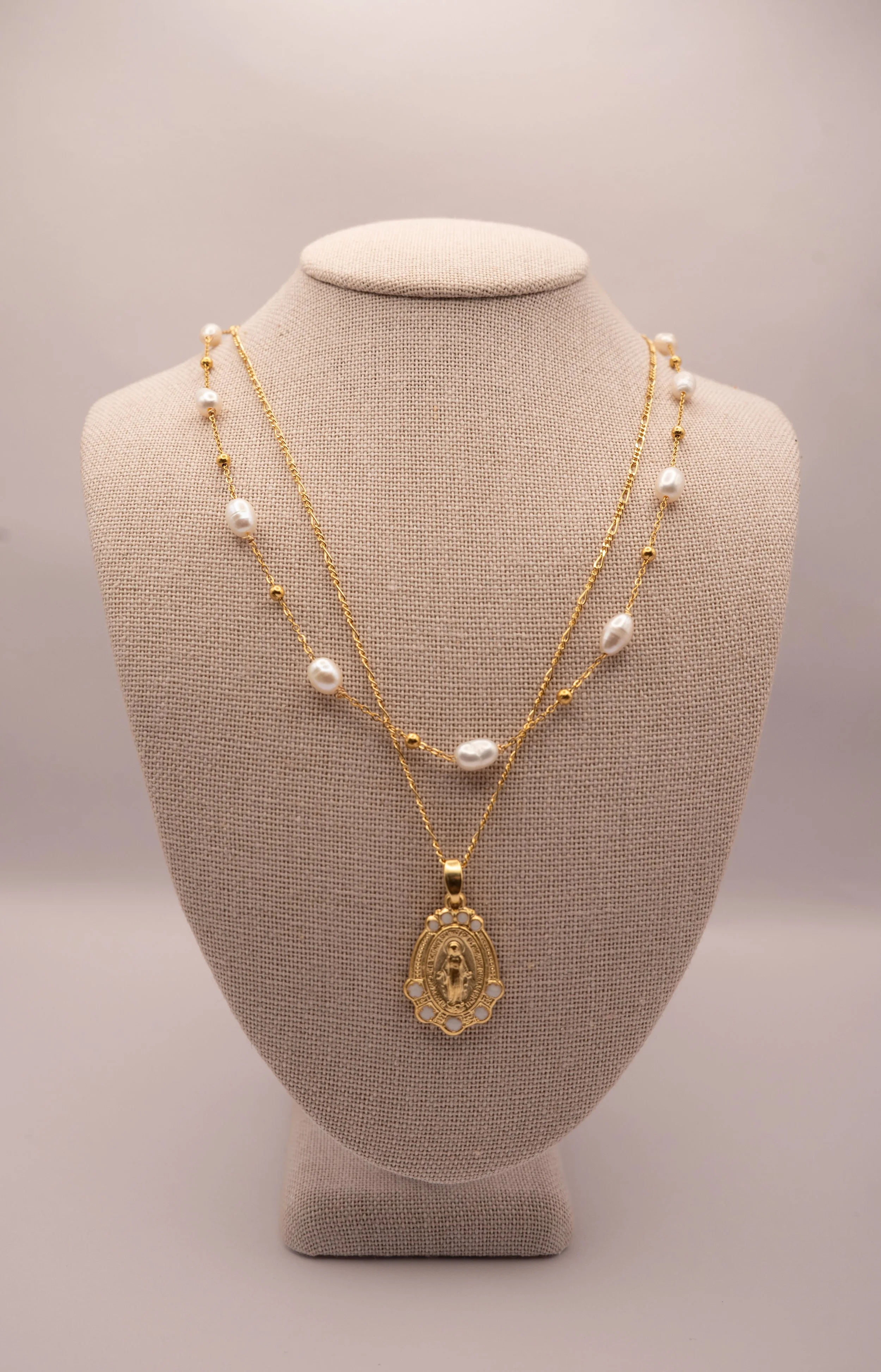 Two layered gold necklaces with white pearls displayed on a beige jewelry stand, with a religious pendant hanging from the bottom necklace.