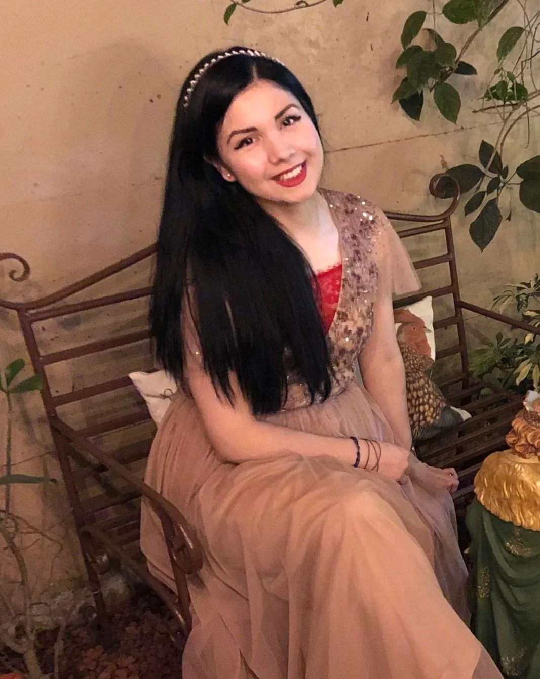Young woman sitting on a wooden bench outdoors, wearing a beige dress with glitter details, smiling, with long black hair and a headband, surrounded by greenery and decorative pillows.