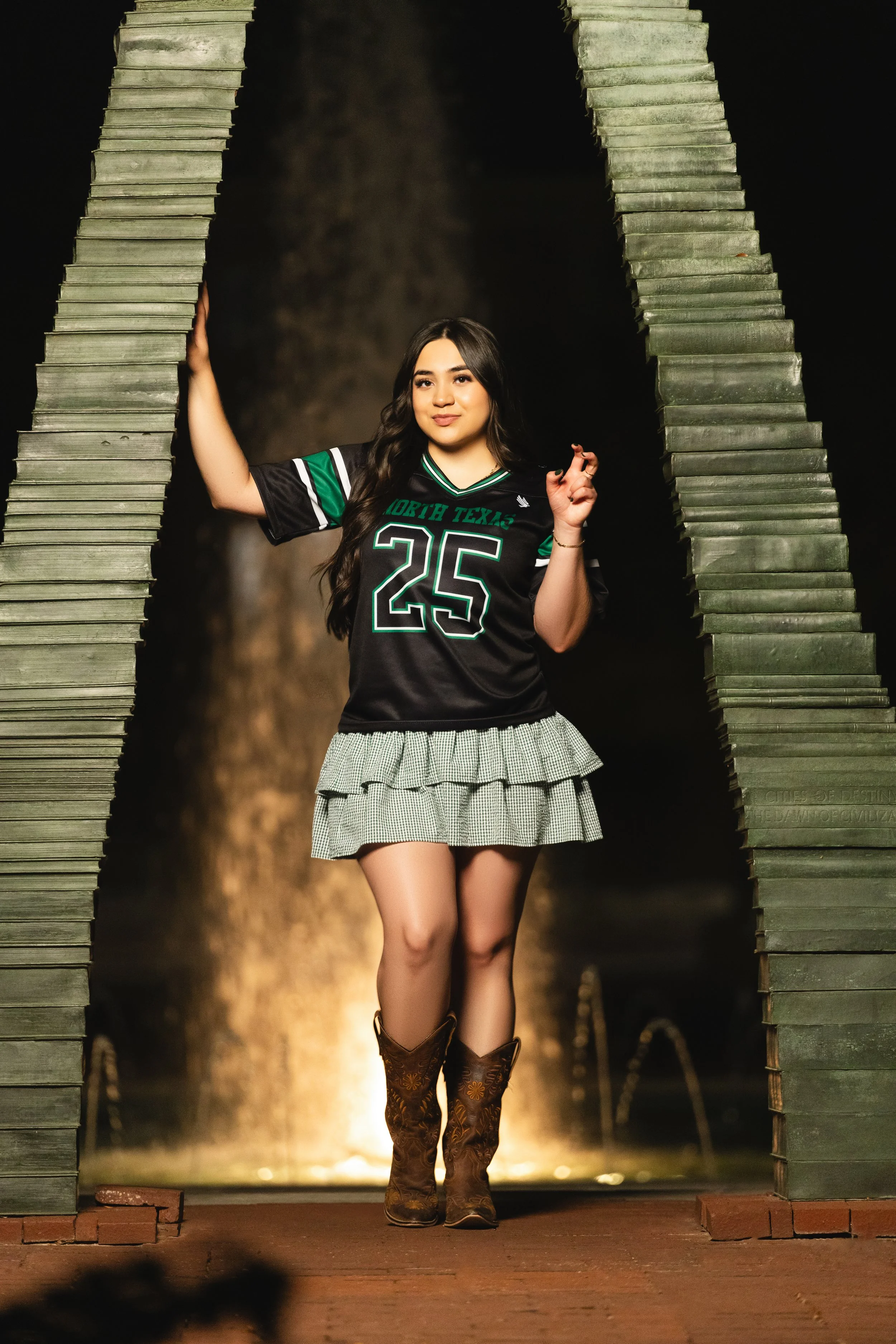Young woman standing outdoors at night, wearing a black sports jersey with the number 25 and the words 'North Texas,' a ruffled skirt, and cowboy boots, holding onto a large wooden archway. photographed by Soba Grad, a graduation photography studio