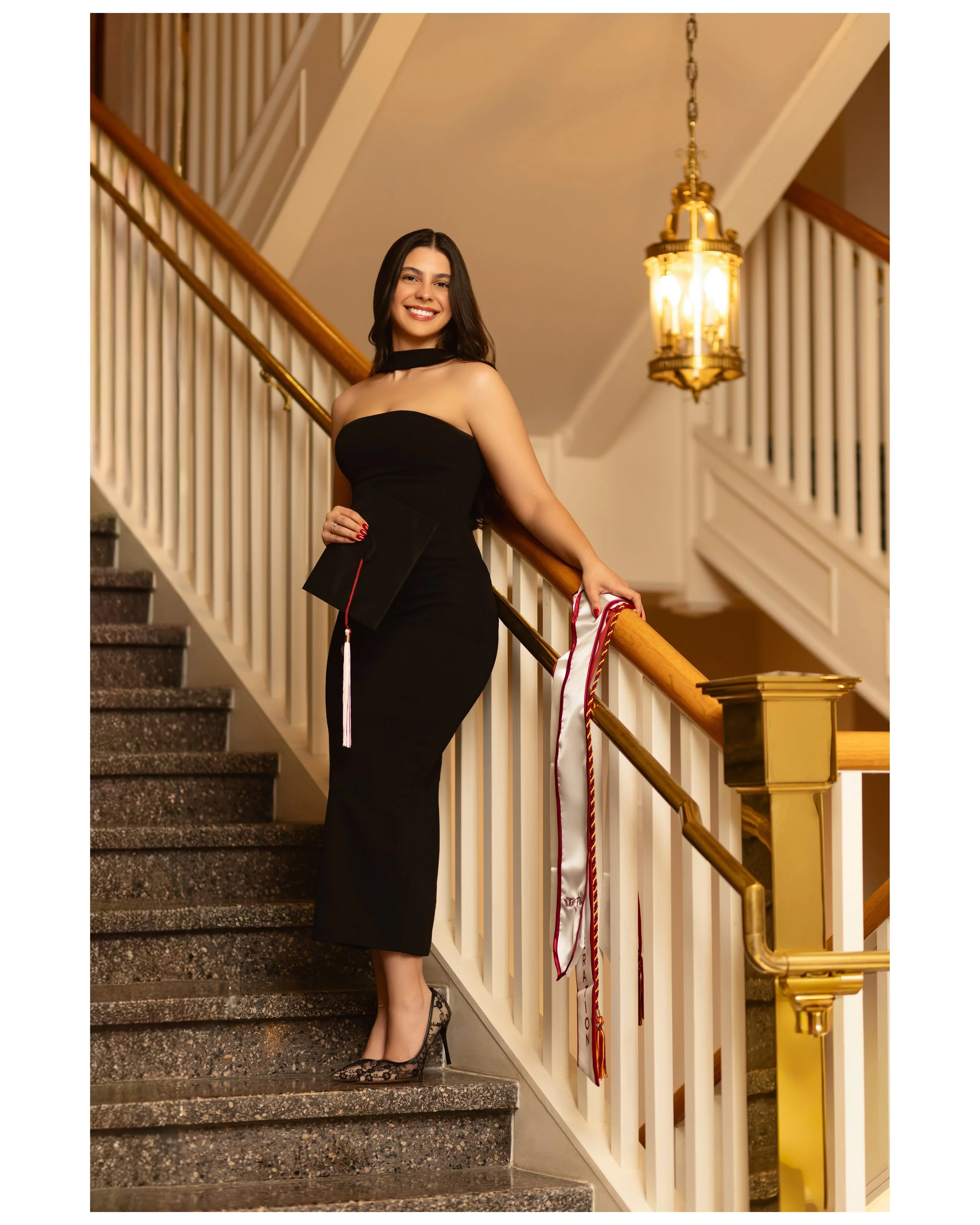 A woman in a black strapless dress and heels is standing on a staircase, holding a graduation cap and wearing a graduation stole, smiling for the camera in an elegant interior setting. photographed by Soba Grad, a graduation photography studio.