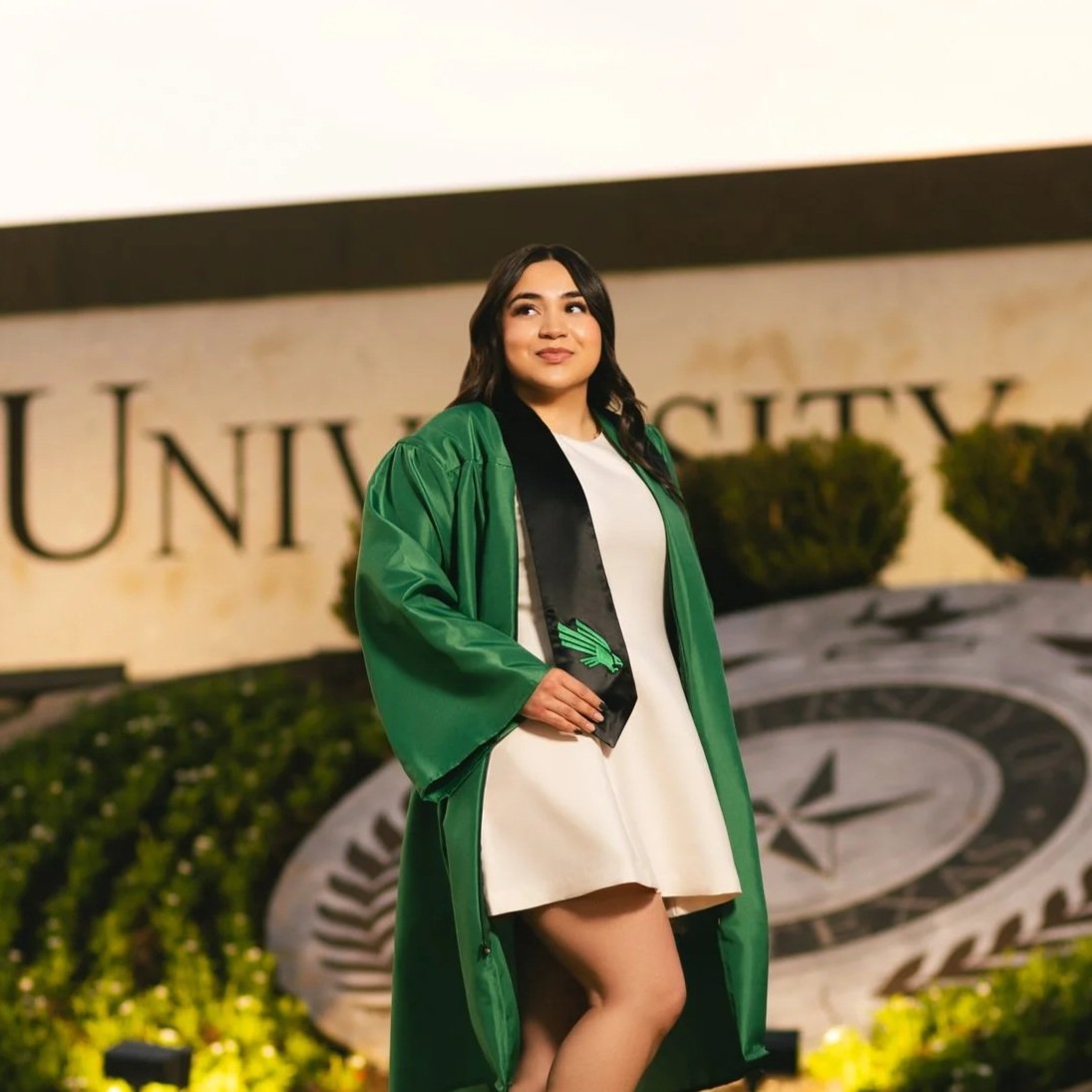 Young woman in a white dress and green graduation gown standing outdoors in front of a university sign. photographed by Soba Grad, a graduation photography studio in Dallas, Texas