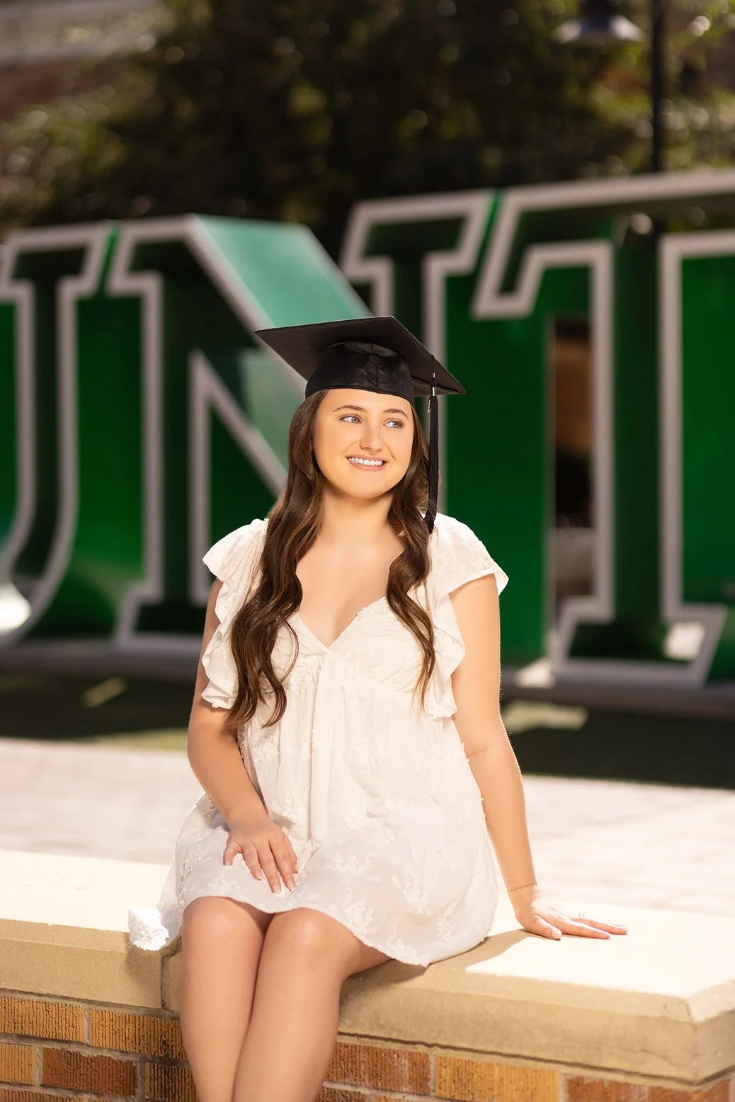 A young woman in a white dress and black graduation cap sitting on a ledge during graduation day outdoors.