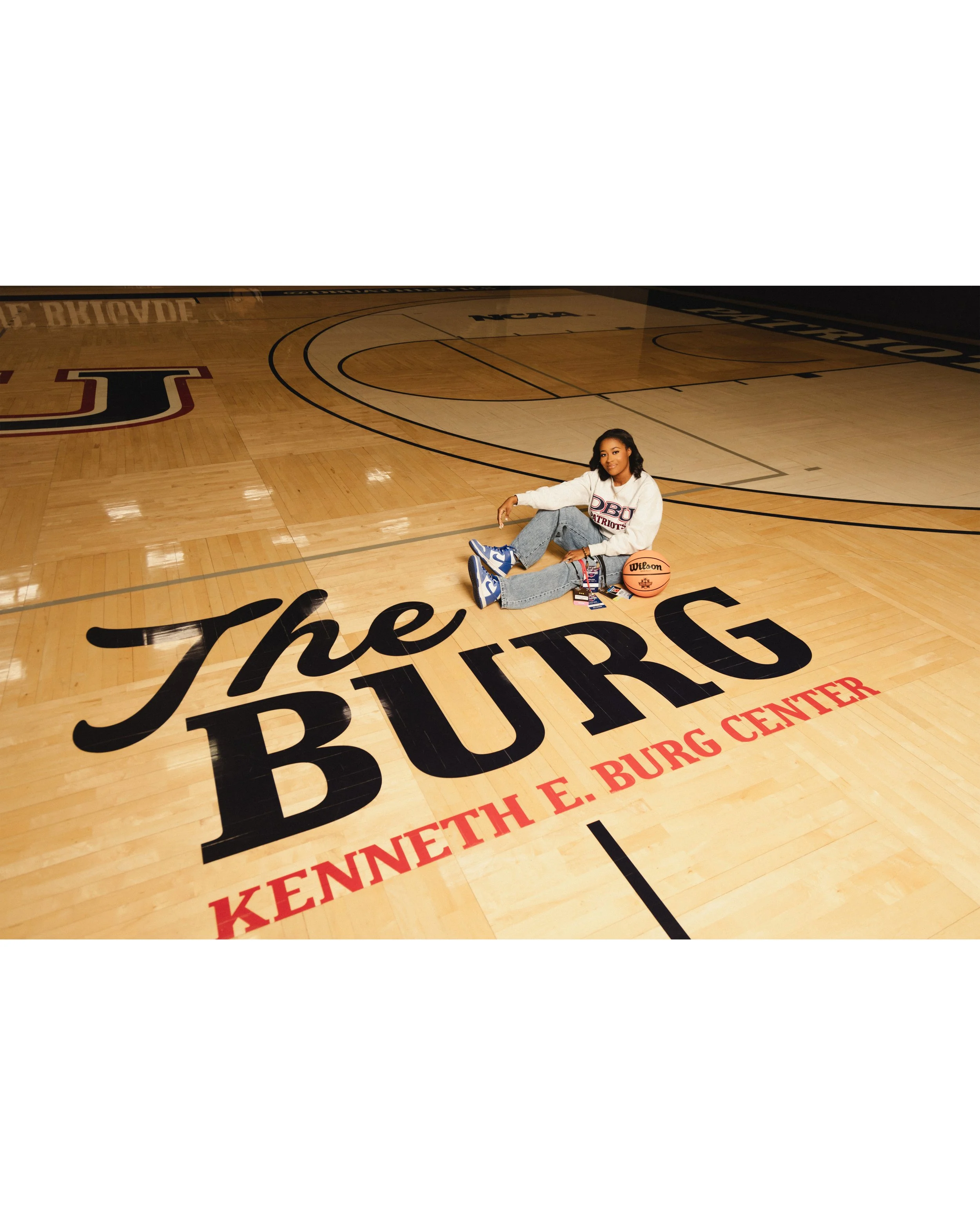 A woman sitting on a basketball court with a basketball, tennis shoes, and a sweatshirt that says "DBU Patriots." The court features a large, colorful inscription that reads "The Burg" and "Kenneth E. Burg Center." photographed by Soba Grad.